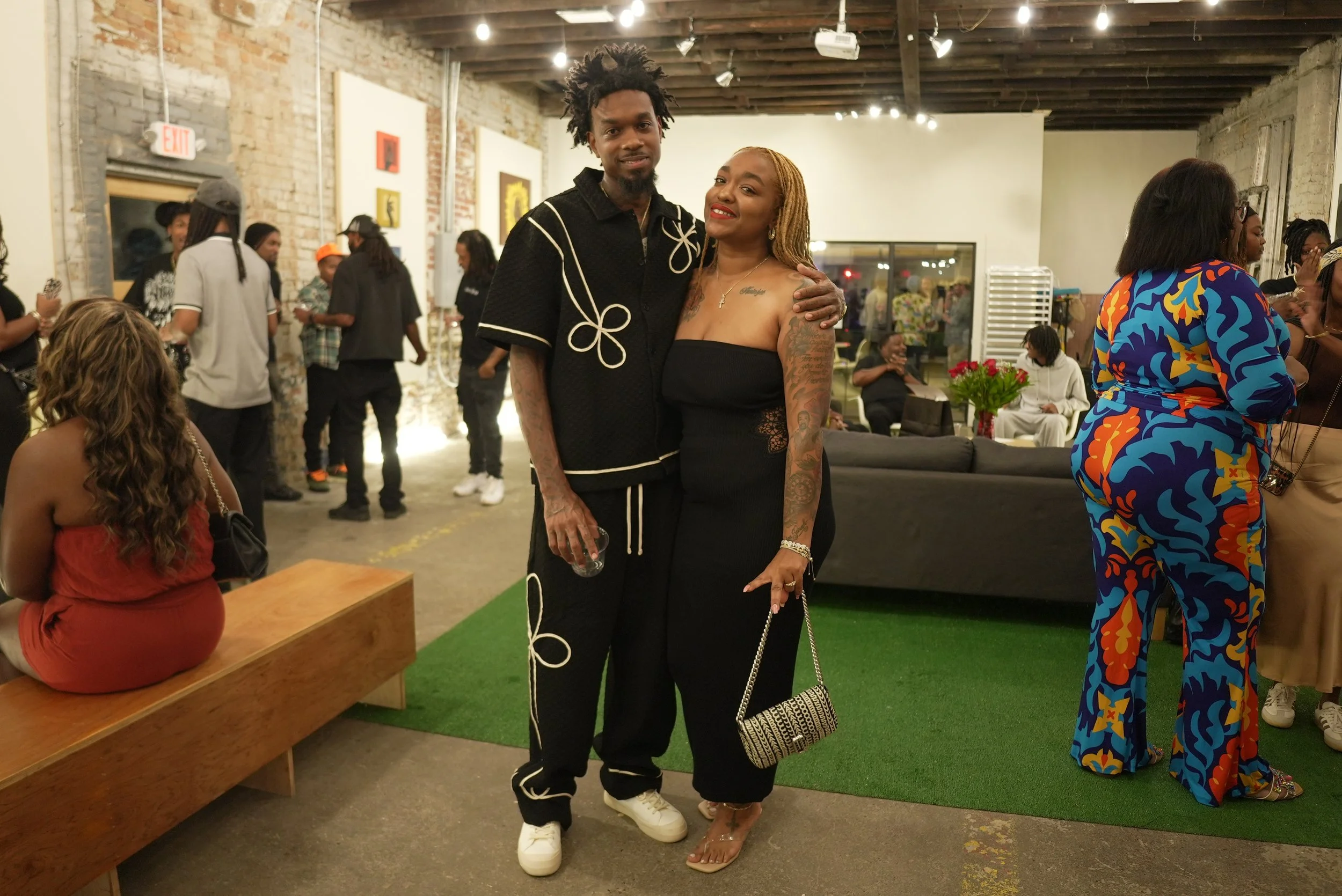Two people standing in an art gallery, smiling at the camera. The man on the left has curly hair, a beard, and tattoos on his arms, wearing a black shirt and pants with white accents. The woman on the right has blonde hair, tattoos on her chest and a