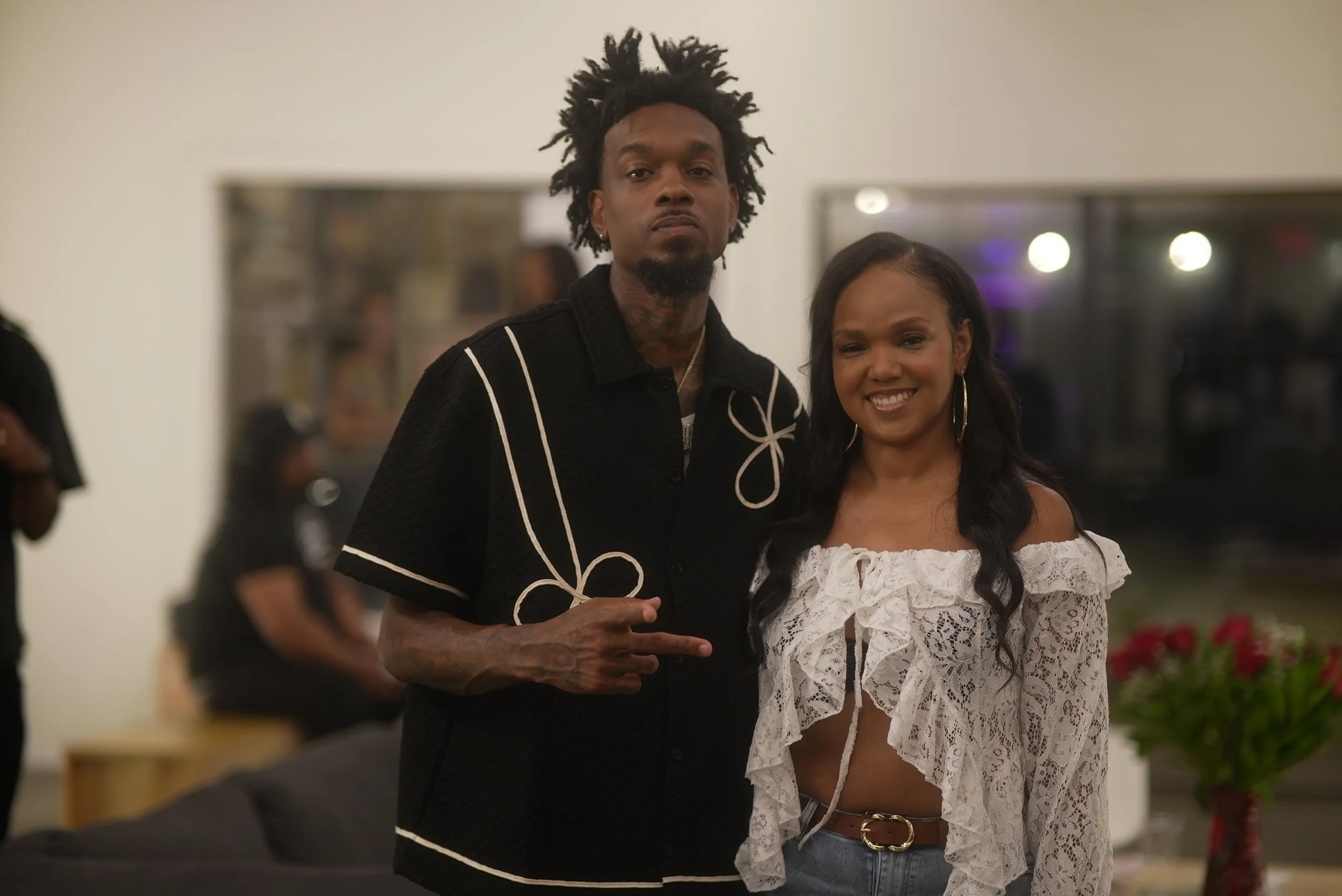Two people, a man and a woman, standing together indoors. The man has dark, curly, spiky hair and a goatee. The woman has long dark hair and is smiling, wearing a white lace off-shoulder top and hoop earrings.