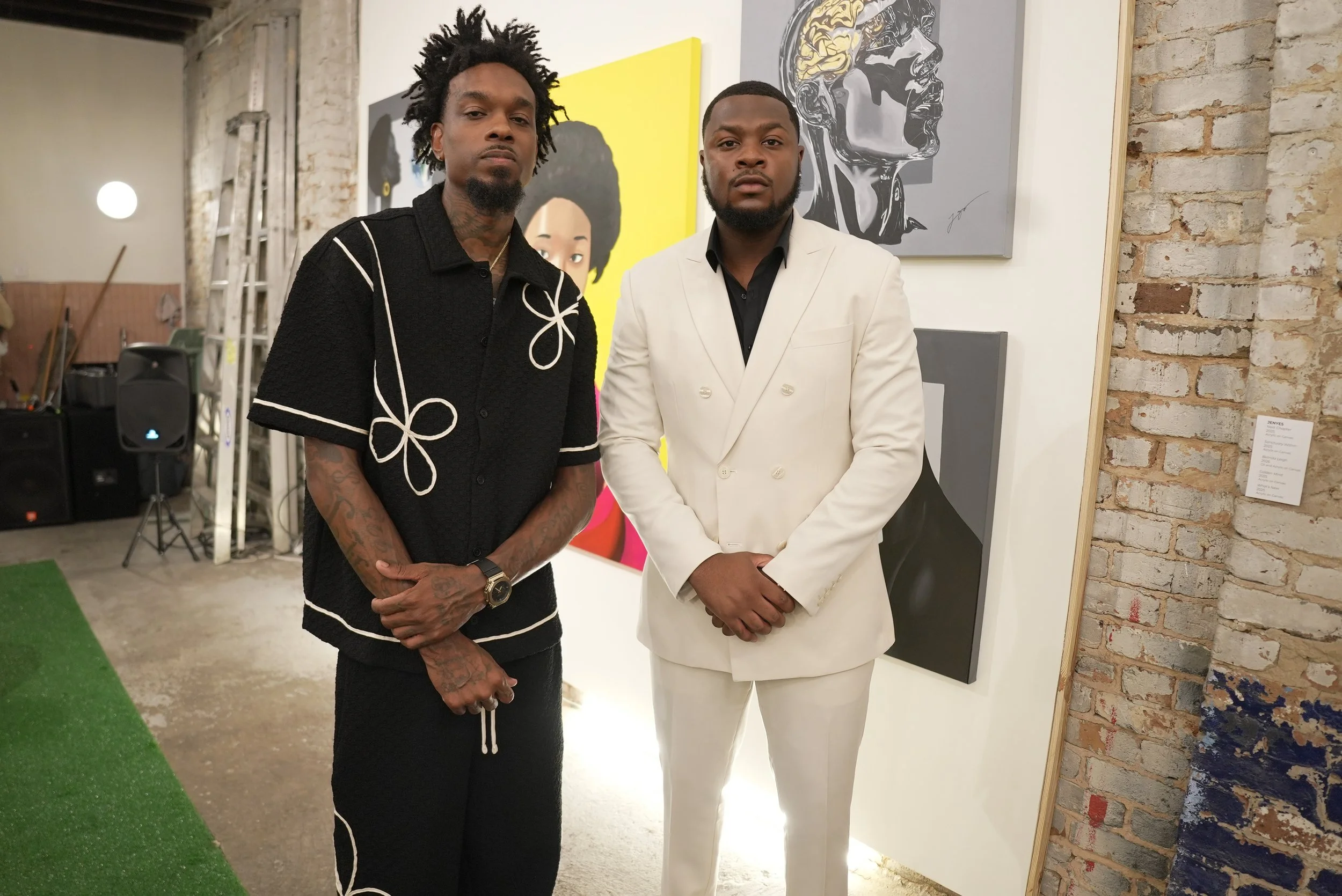 Two men standing side by side in an art gallery with paintings behind them, one wearing a black shirt with white embroidery and the other in a white suit.
