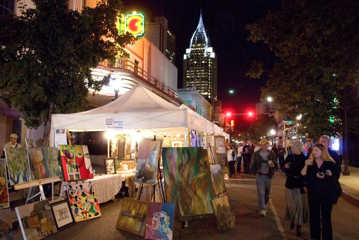 Night street art festival with paintings displayed on stands and easels outside a tent, with people walking and chatting under streetlights, and a tall illuminated building in the background.