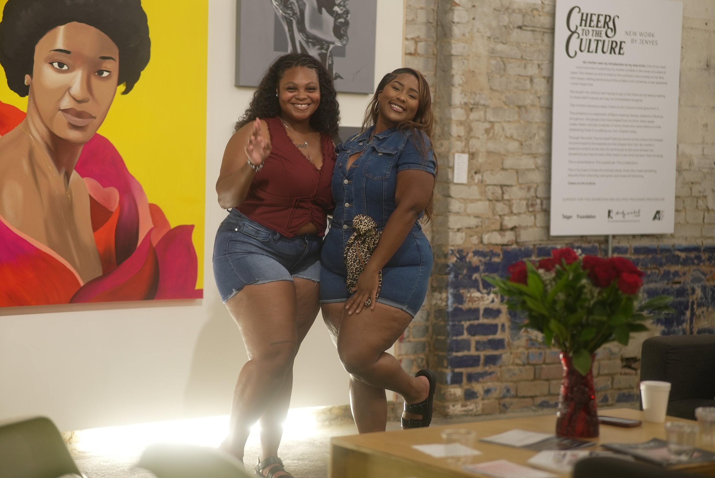 Two women smiling and posing together at an art exhibition, standing in front of colorful artwork and a brick wall, with a table and flowers in the foreground.