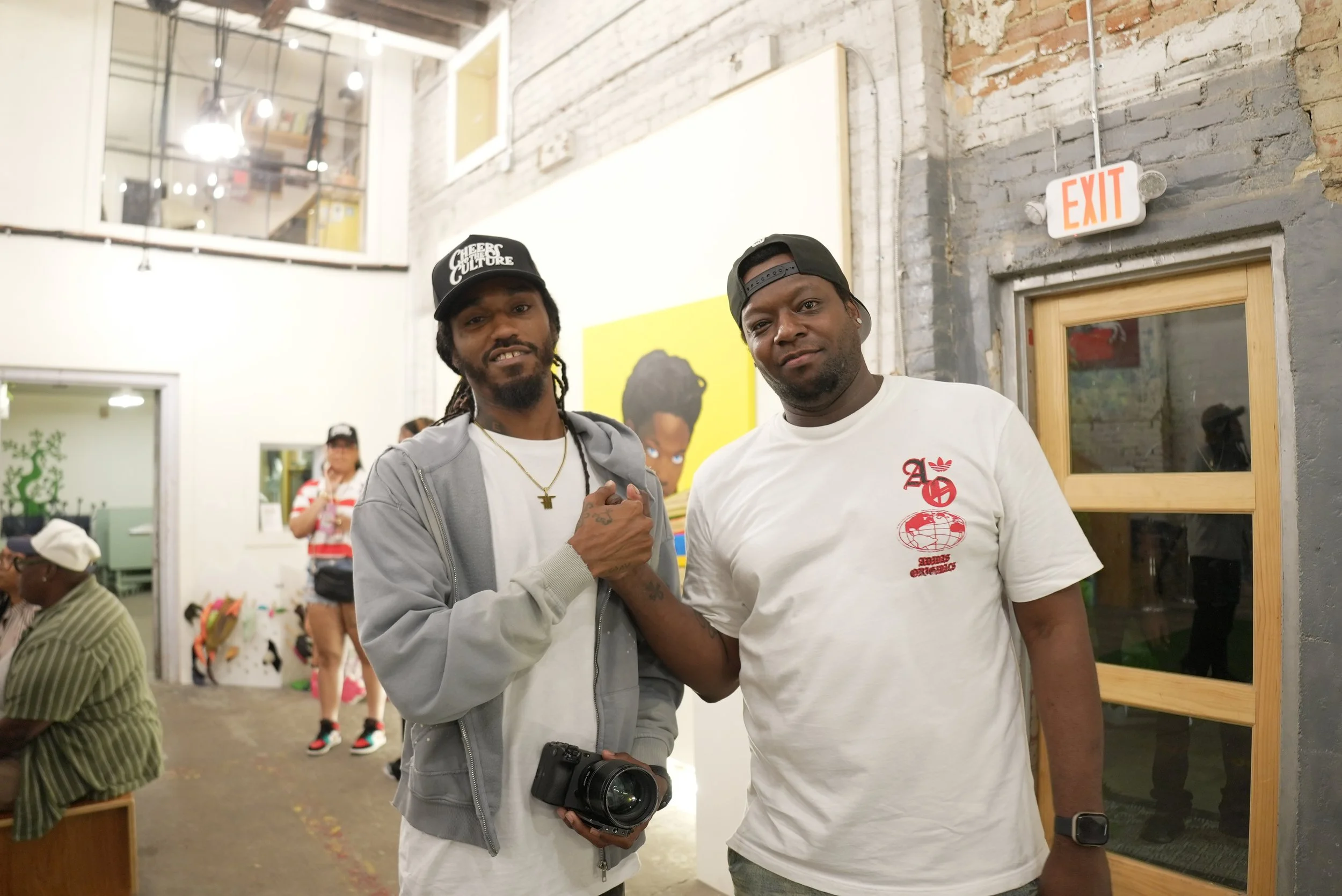 Two men with dark skin standing together at an indoor event, shaking hands and smiling at the camera. The man on the left has dreadlocks, is wearing a black cap with white text, a gold chain, and a gray hoodie. The man on the right has a black cap ba