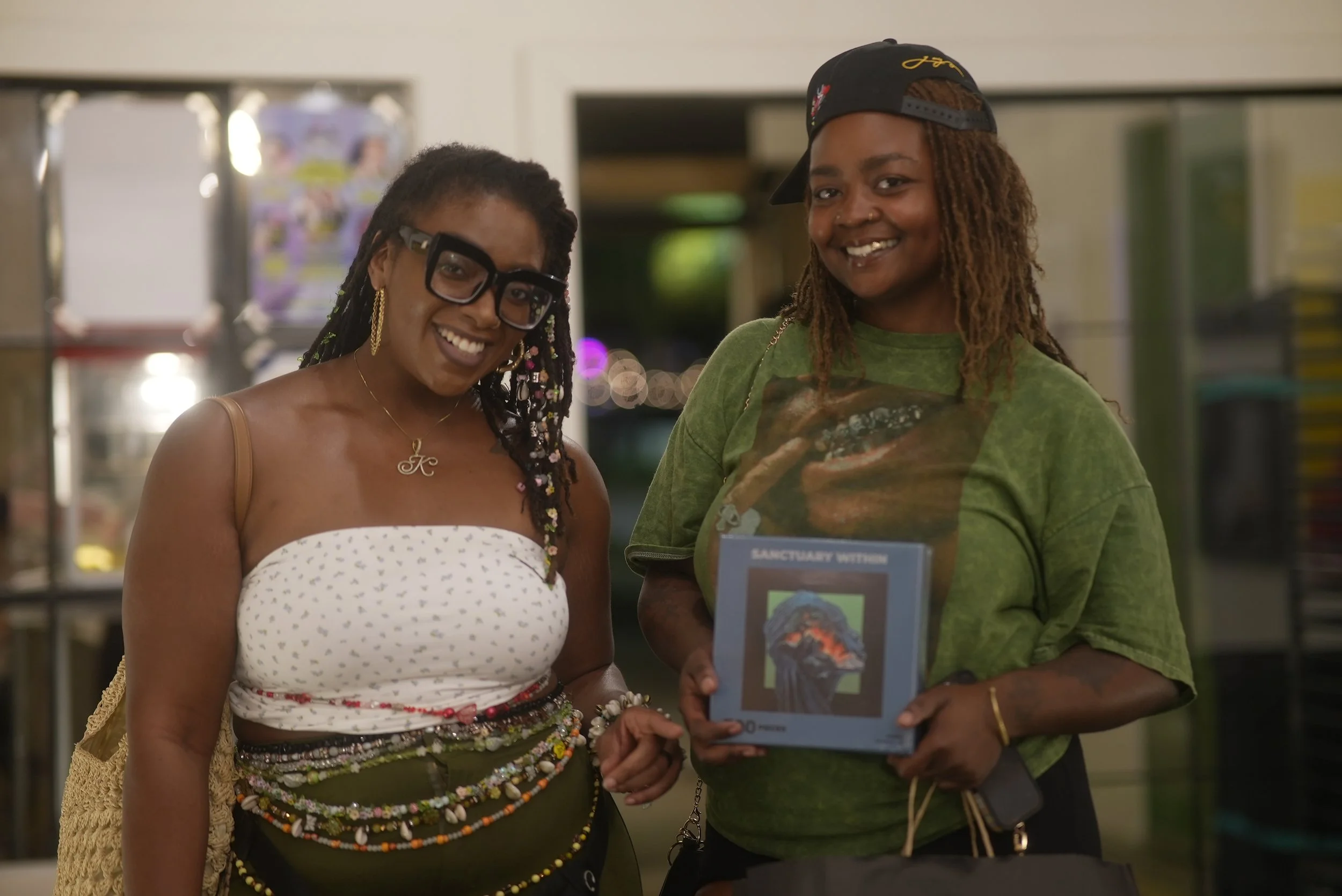 Two women smiling, one in a white dress with colorful beaded jewelry and glasses, and the other in a green shirt with a black cap worn backwards, standing inside a store.