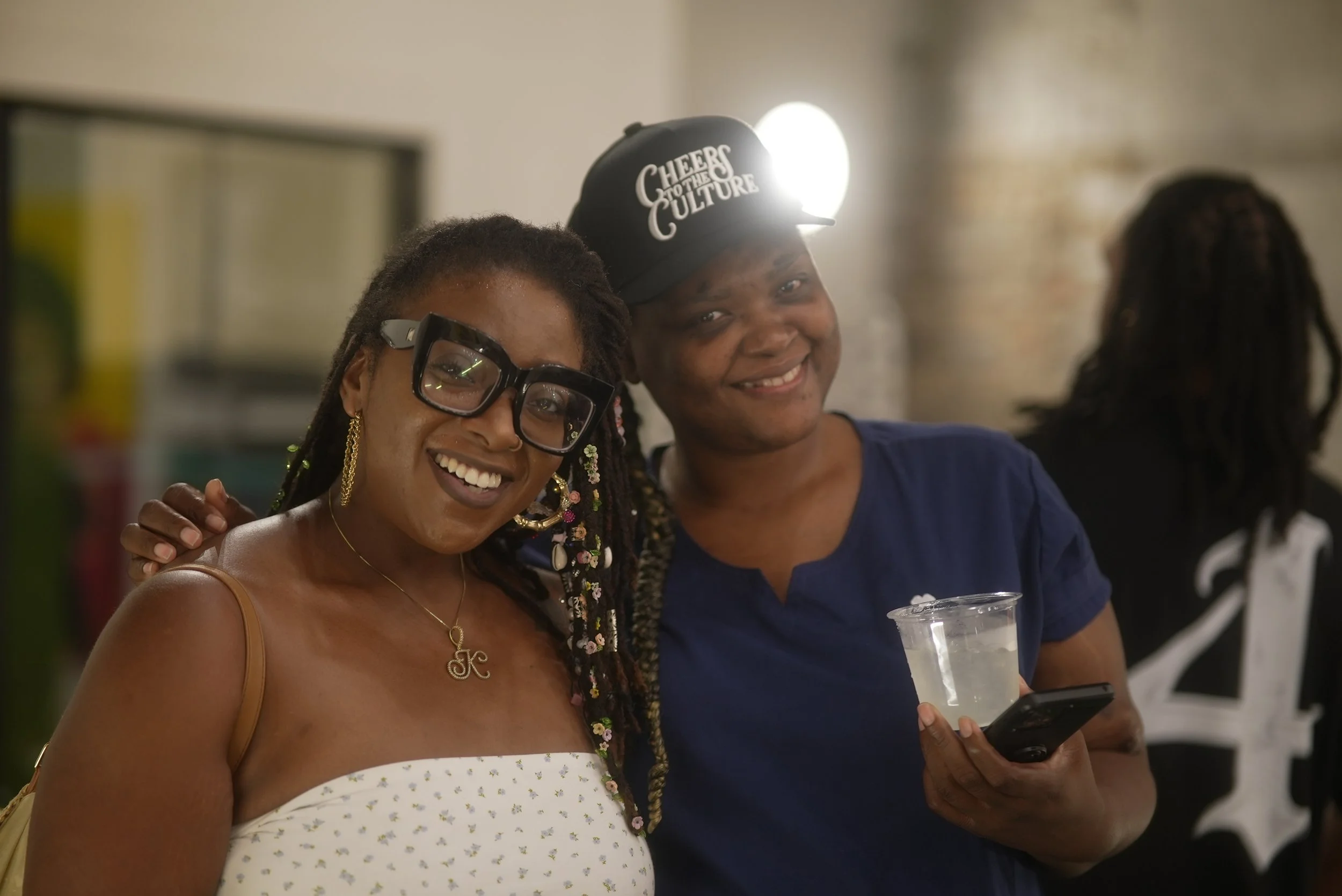 Two women smiling, one holding a drink and a phone, the other wearing glasses and a white dress with small floral pattern, in an indoor setting with a blurred background.