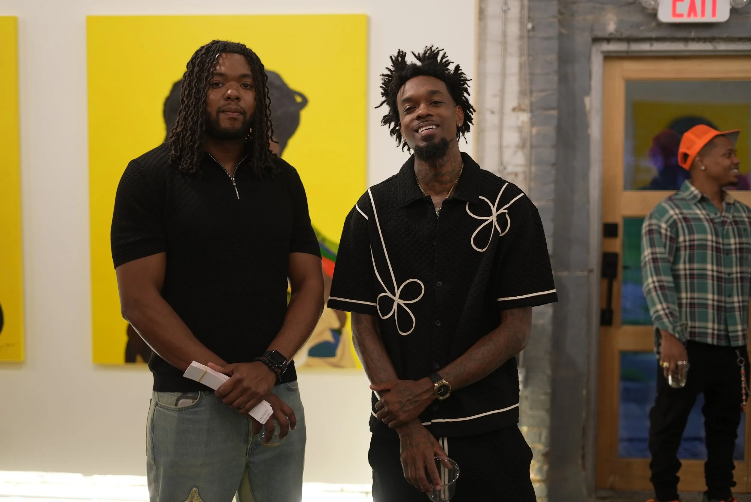 Two men standing side by side in an art gallery, with one man holding a piece of paper and the other smiling, with artwork on the wall behind them.
