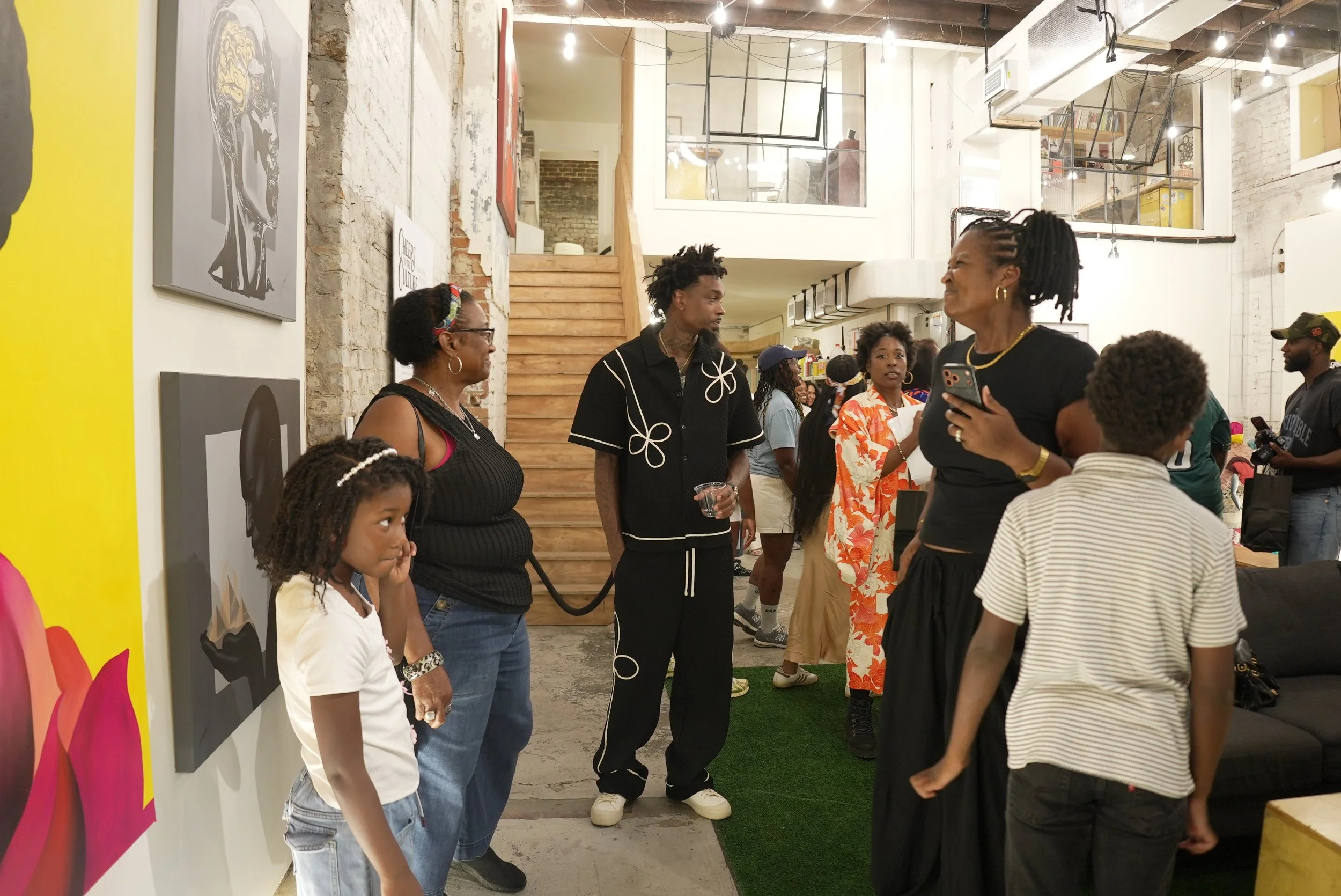 A group of diverse people, including women and children, are gathered inside an art gallery or exhibition space. The setting features exposed brick walls, wooden stairs, and modern lighting. Some individuals are engaging in conversation, while others