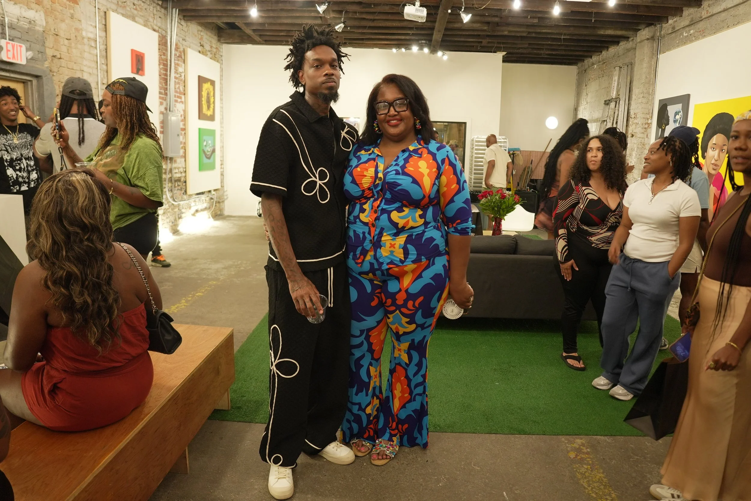 Two people stand close together in a gallery space, smiling at the camera. The person on the left has dark skin, dreadlocked hair, and is wearing a black shirt with white piping and black pants. The person on the right has dark skin, black hair, and 