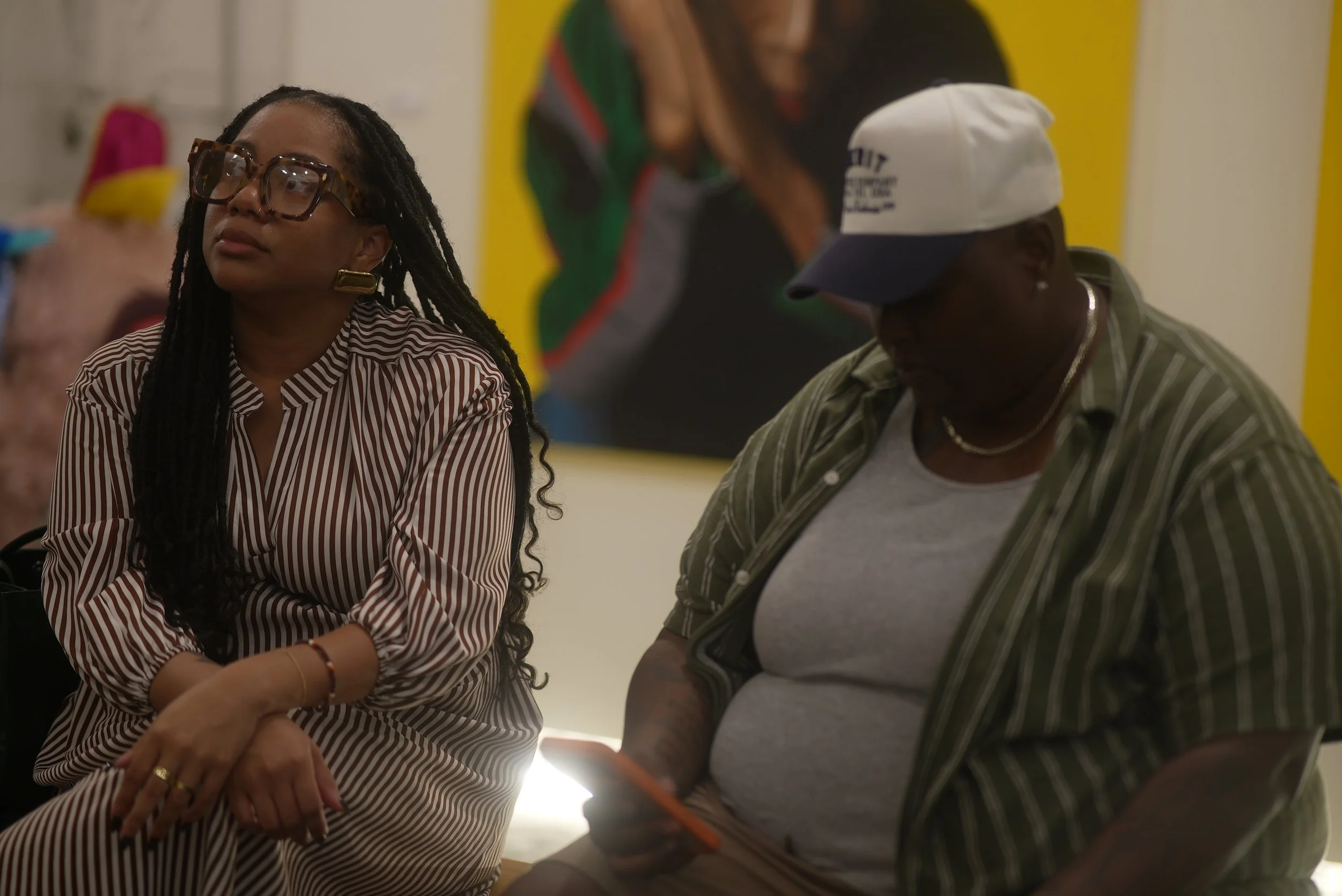 Two women sitting side by side, one with medium skin and braided hair wearing glasses and a striped shirt, the other with dark skin wearing a white cap, striped shirt, and jewelry, in an indoor setting with a yellow and colorful background.