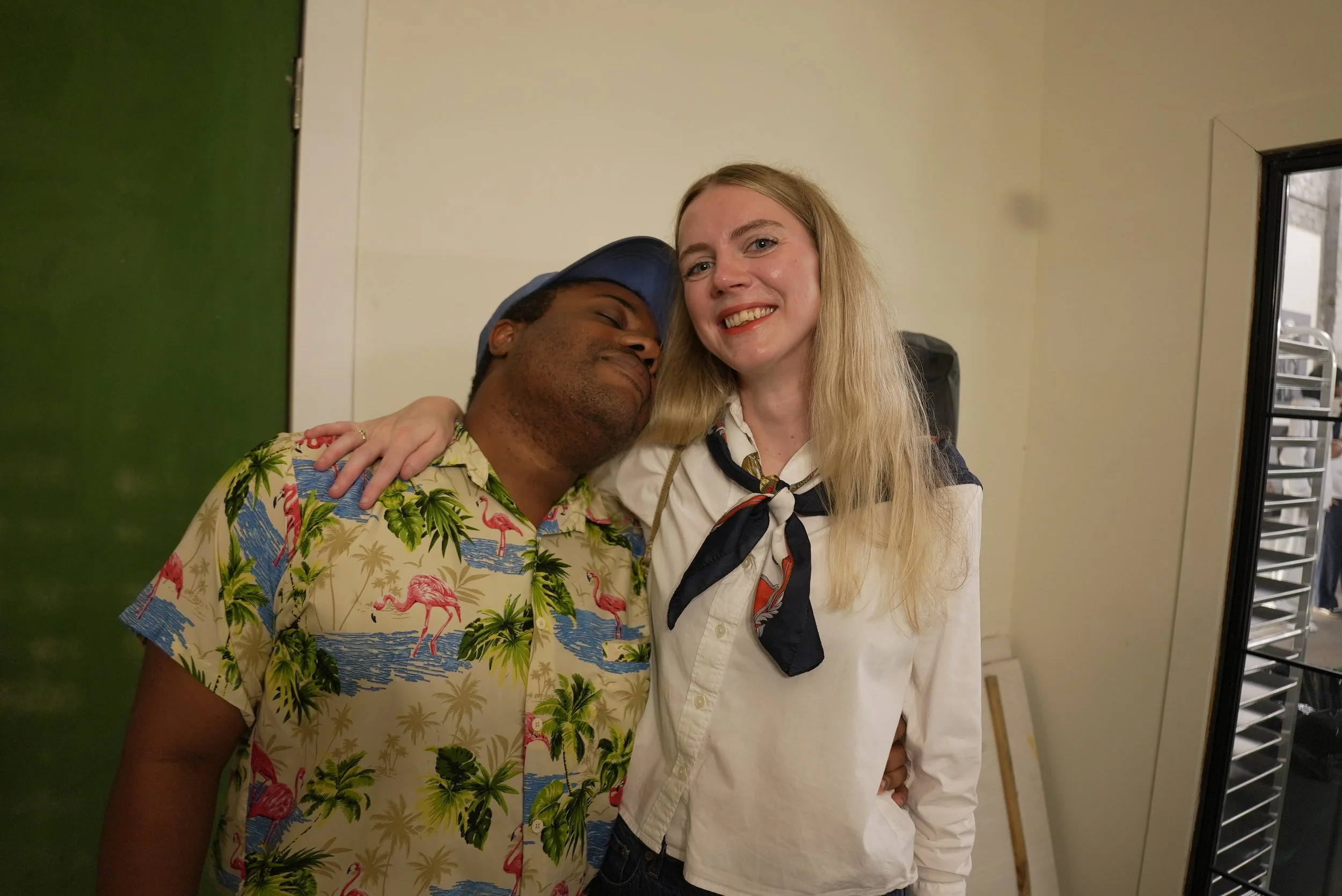 A man and a woman are hugging and smiling at the camera inside a room with light-colored walls and a black-framed window, with the woman wearing a dark necktie and the man wearing a colorful shirt with flamingos and palm trees.
