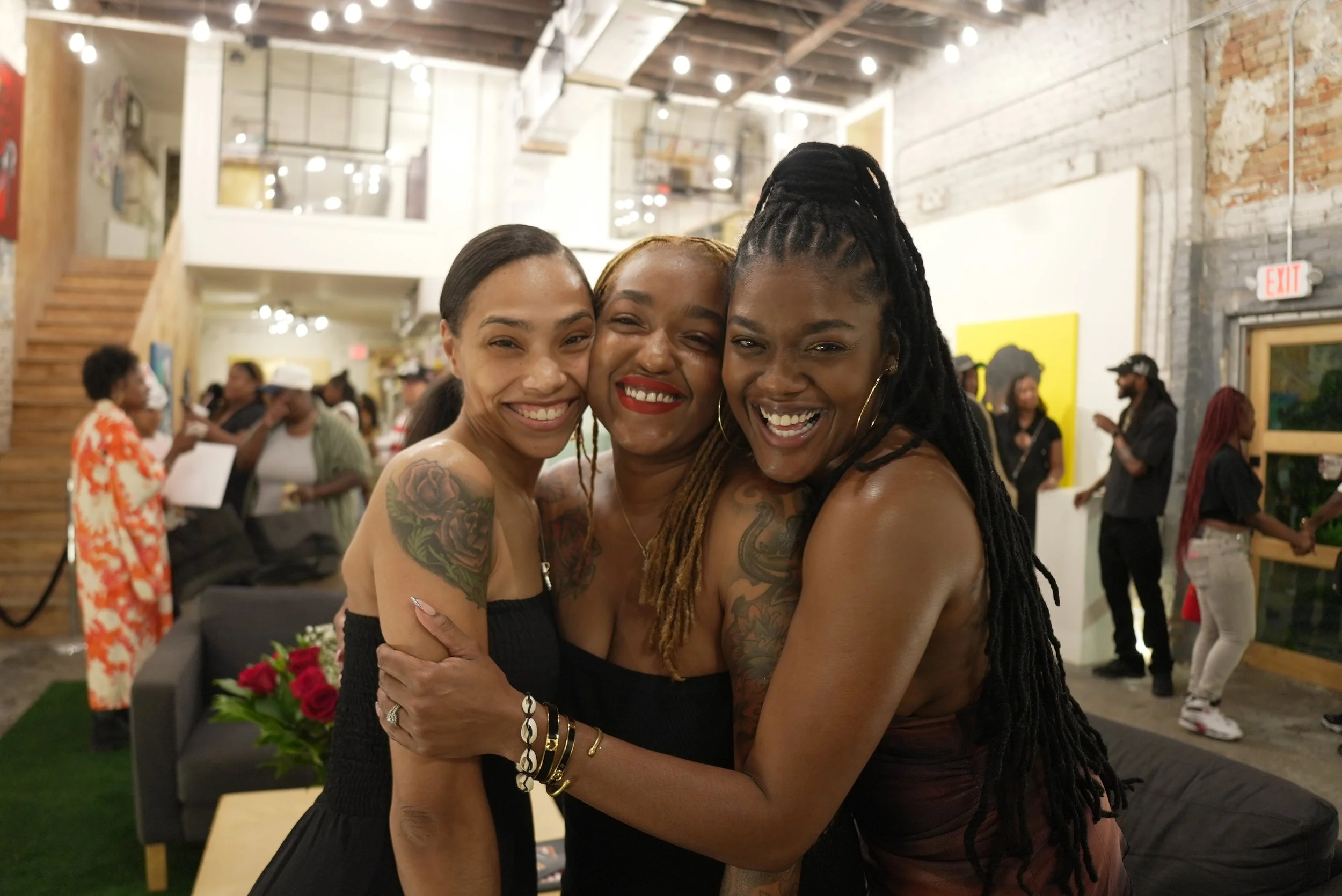 Three women embracing and smiling inside an art gallery or studio space with exposed brick walls and wooden ceiling beams.