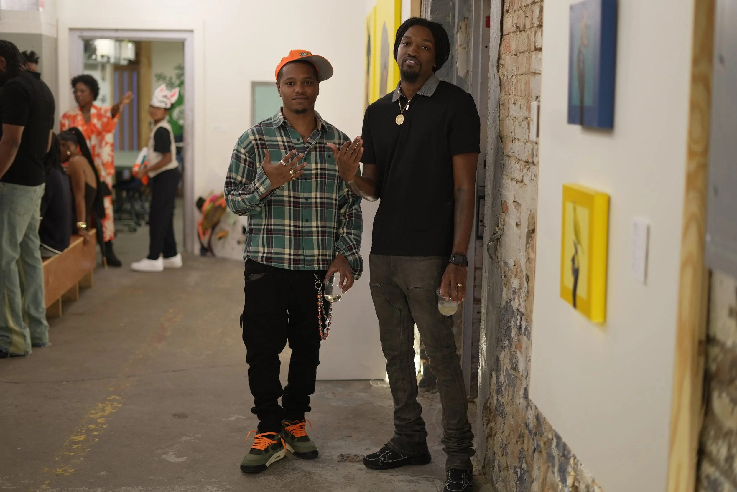 Two men stand side by side in an art gallery, leaning against a brick wall. The man on the left wears a plaid shirt, orange cap, and holds a wine glass and rosary beads. The man on the right wears a black polo shirt, gold chain, and also holds a wine