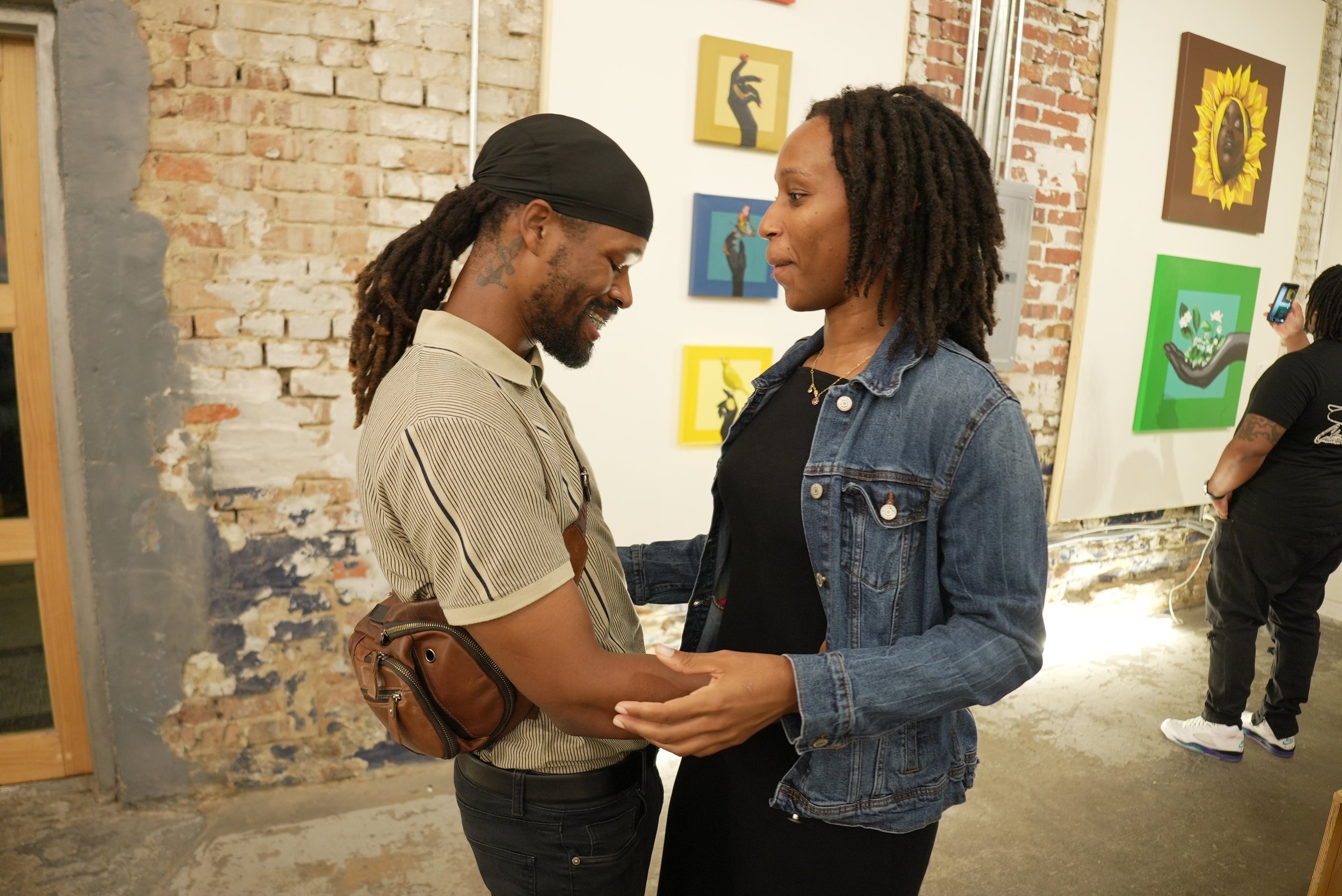 A man and a woman with dreadlocks are standing close together, holding hands, and looking into each other's eyes inside an art gallery with brick walls and colorful artwork.