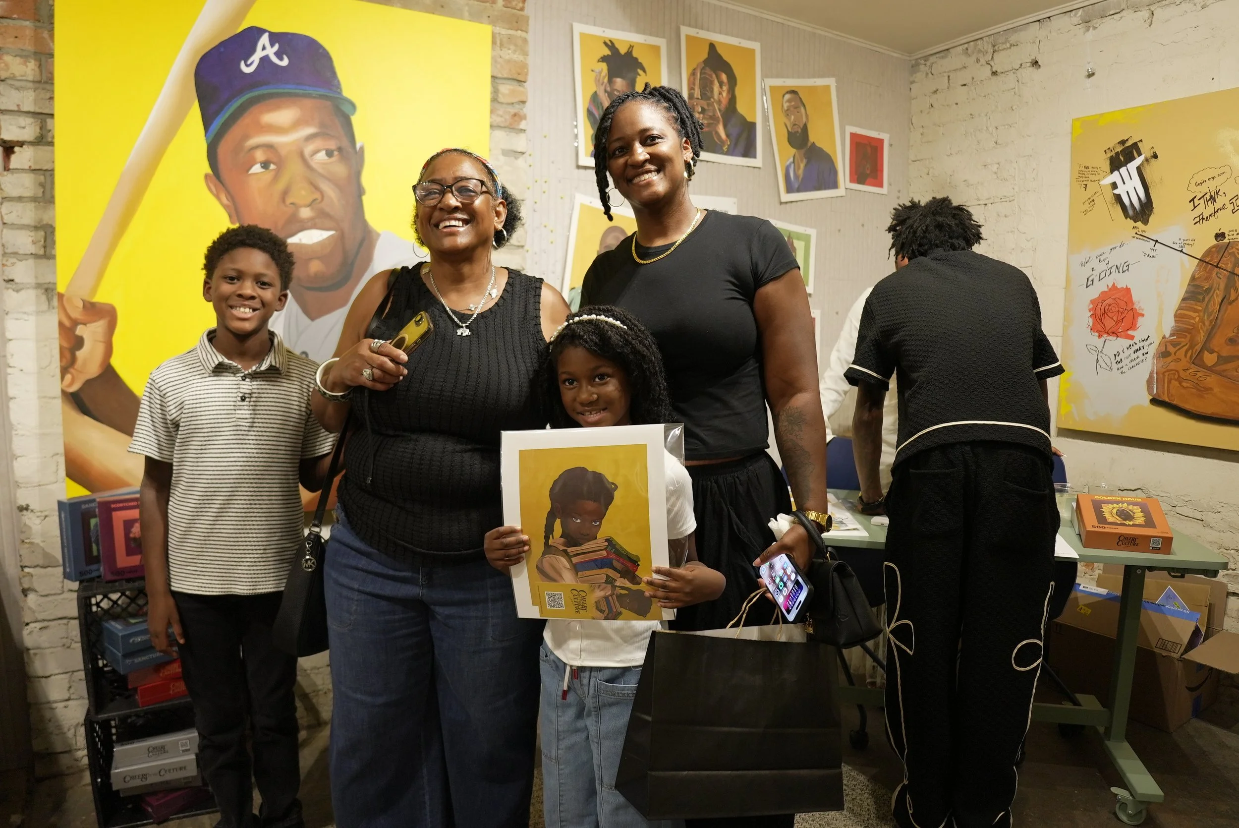 A group of four people, two women and two children, standing in front of artwork with portraits of people on a gallery wall. One woman is holding a poster of a girl with books, and everyone is smiling.