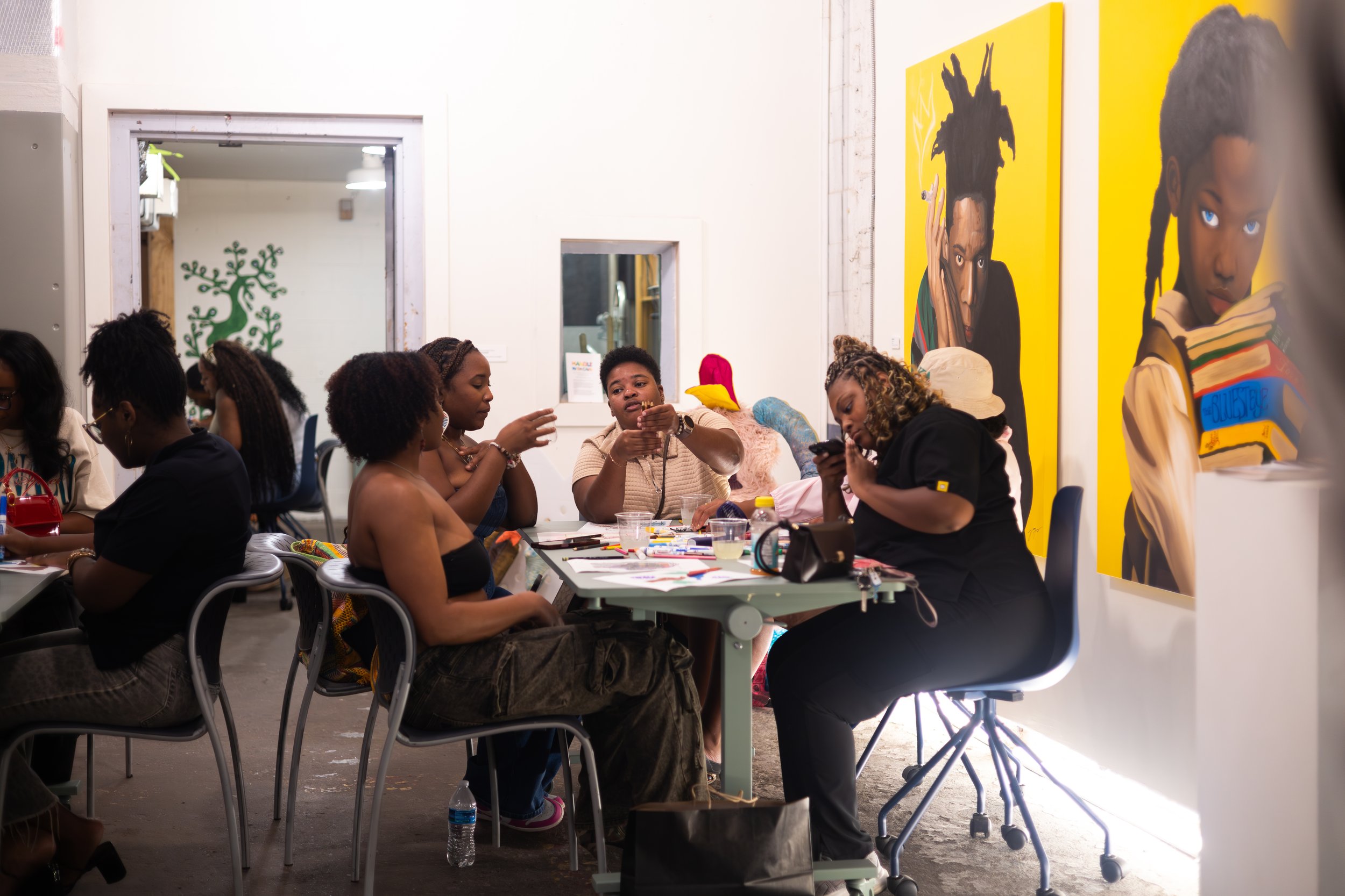 A group of women gathered around a table in an art gallery, looking at artwork and engaging in conversation. The gallery features colorful paintings on the wall and a window with a green tree decal.