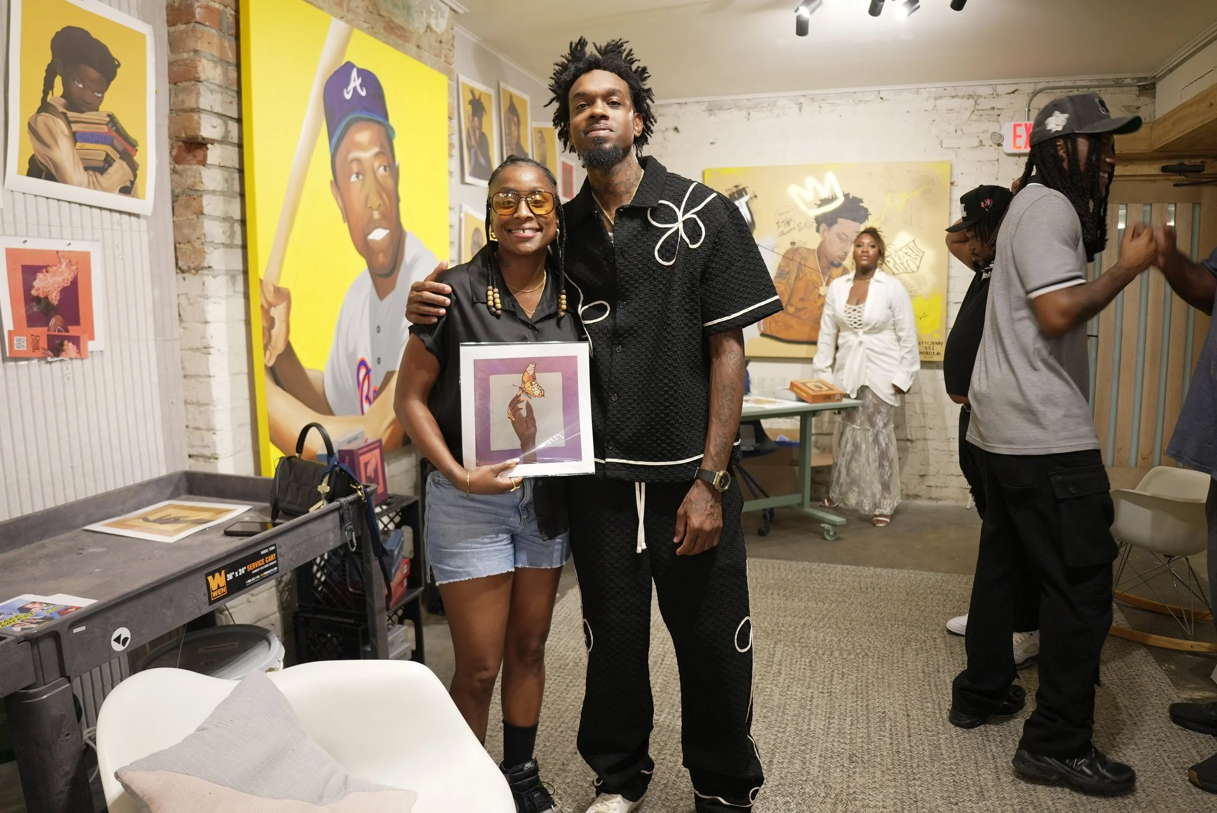 Two people smiling and posing together inside an art gallery, with colorful artwork behind them. The woman on the left has glasses, braided hair, and is holding a framed artwork. The man on the right has dreadlocks and tattoos, wearing a black patter