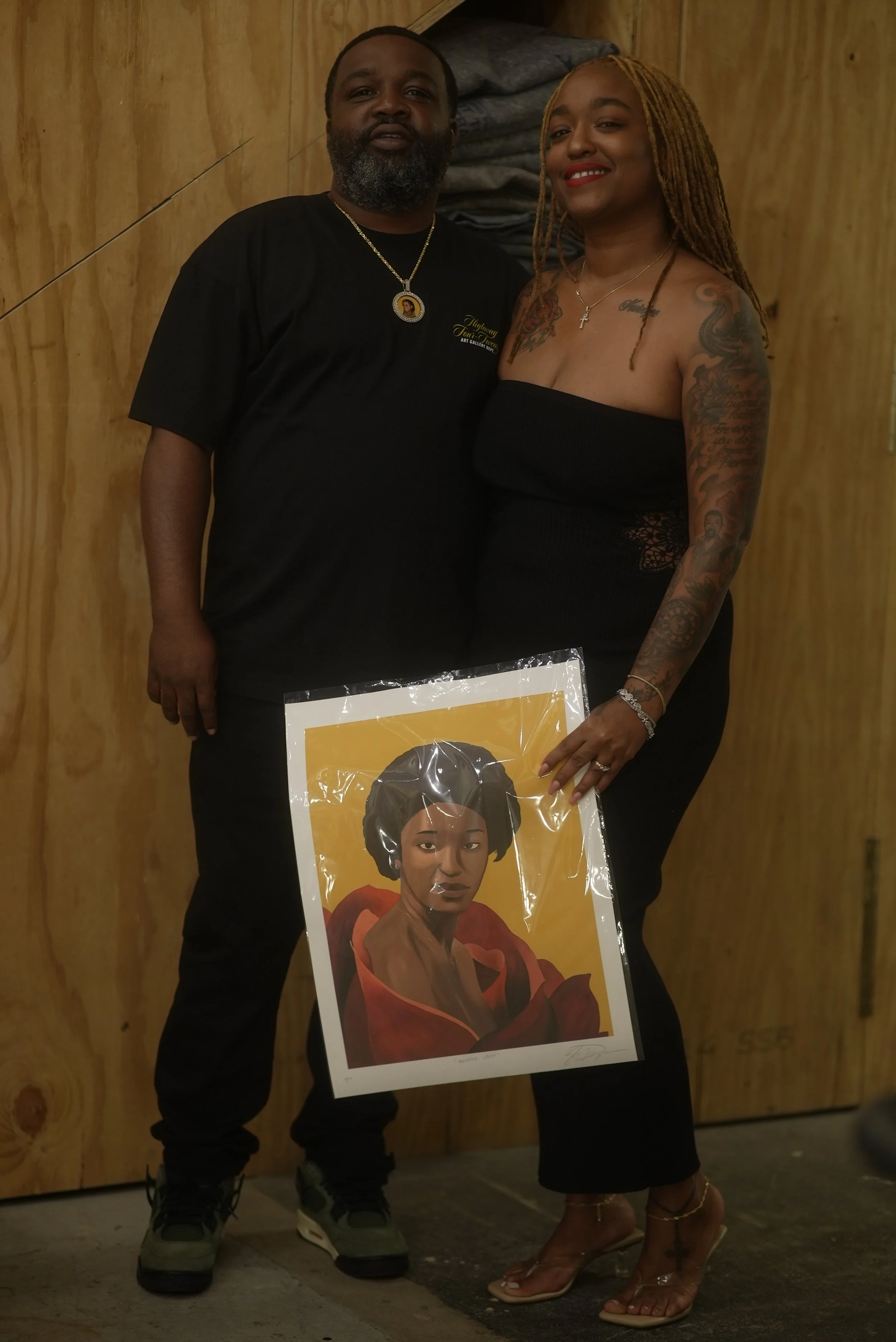 A man and woman standing together indoors, holding a portrait of a woman with natural hair and a confident expression. The man is wearing a black t-shirt with a necklace, and the woman is dressed in a black strapless dress with tattoos on her arms an