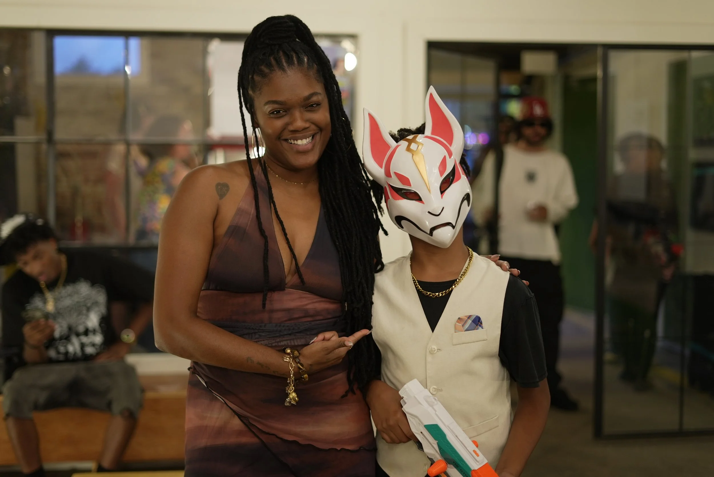 A woman with long dreadlocks and a tattoo on her shoulder smiling with a person wearing a fox mask and a costume. Others are visible in the background in an indoor setting.