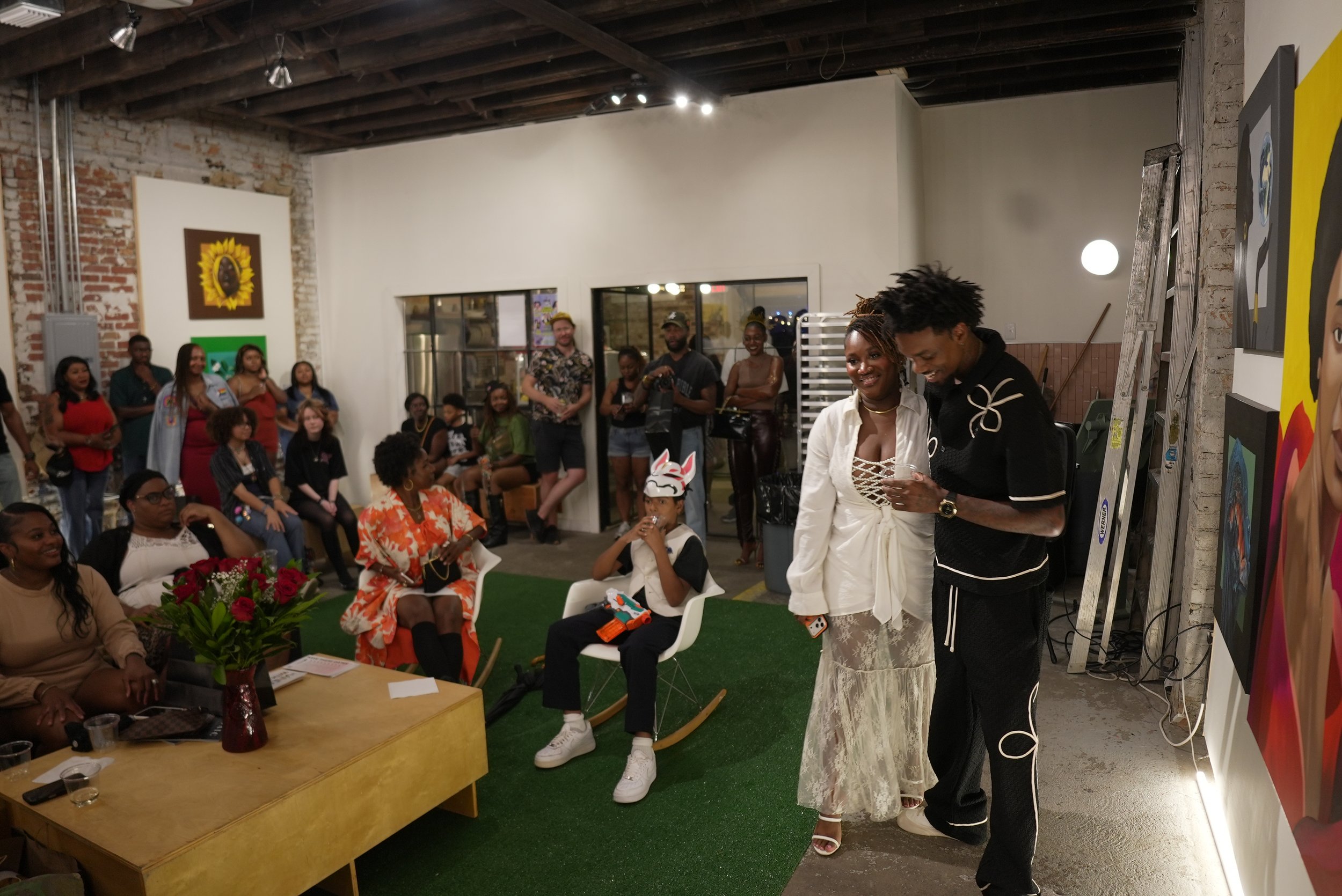 A group of people gathered in an art gallery or studio, with some seated and others standing. Two individuals are at the front, smiling and embracing. Artwork is displayed on the walls, and there is a table with flowers and papers in the foreground.