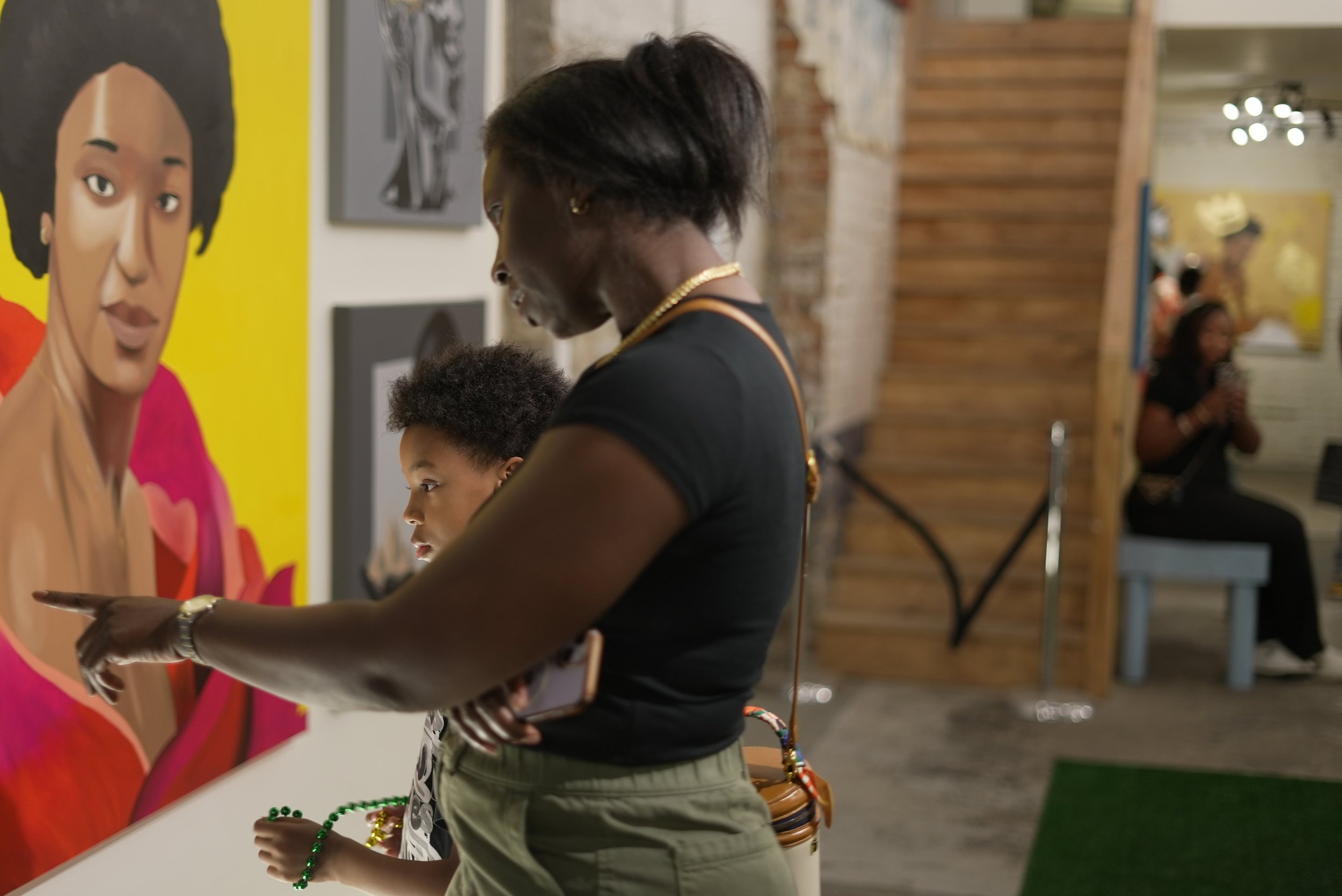 A woman and a child looking at artwork on a gallery wall, in an art gallery with wooden stairs in the background.