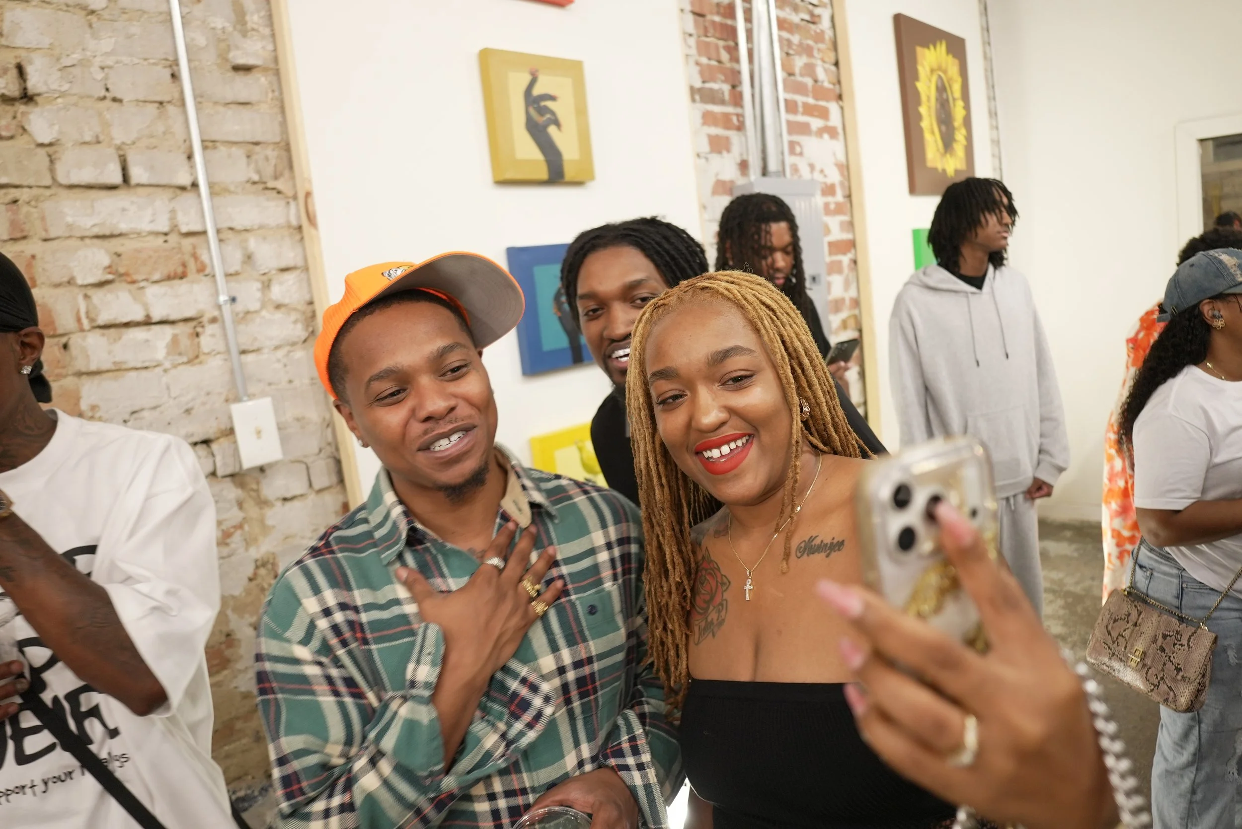 A group of diverse young people at a social gathering, smiling and taking a selfie together in an indoor setting with brick and painted walls, some artwork on the walls.