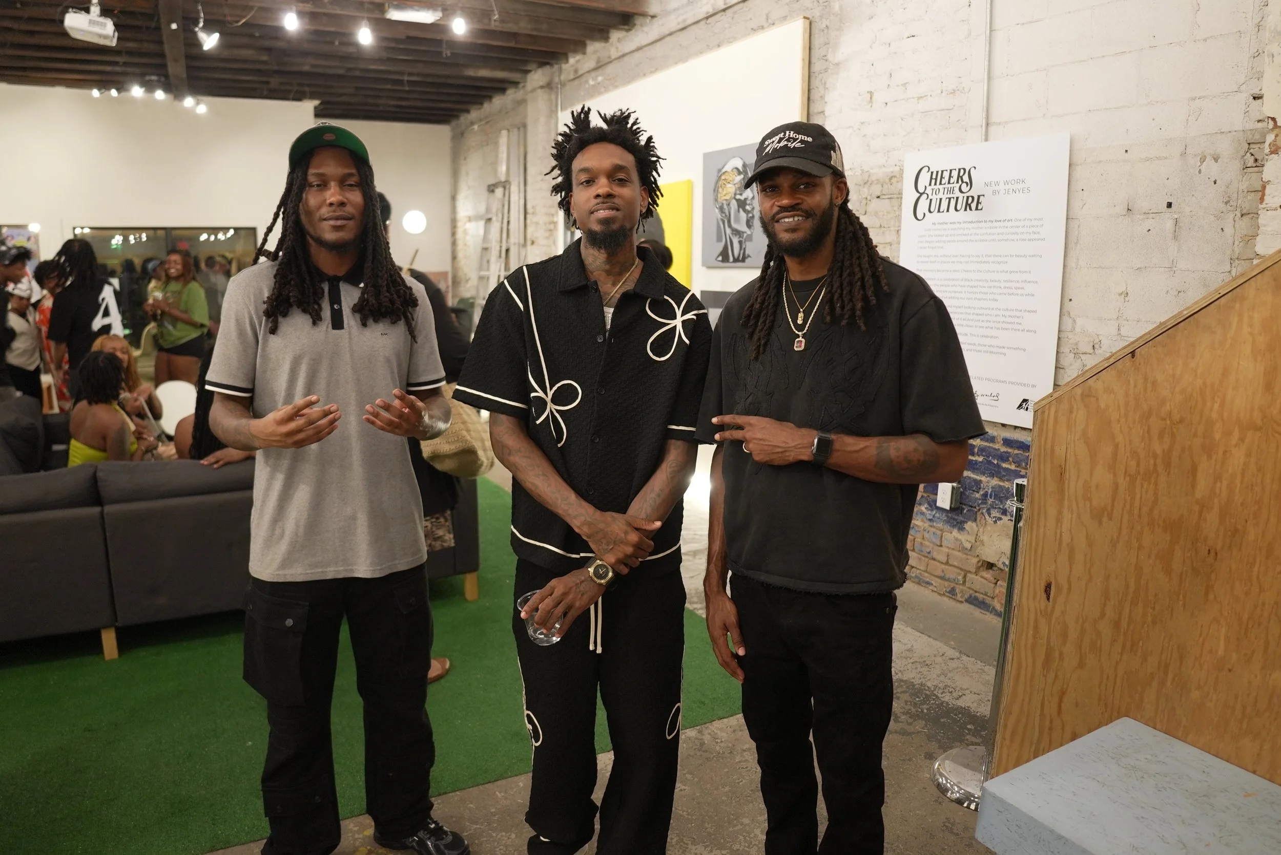 Three men standing together at an art gallery. They are posing for the photo with a white brick wall with paintings behind them. The man on the left is wearing a cap and a gray polo shirt, the man in the middle has curly hair and is wearing a black s