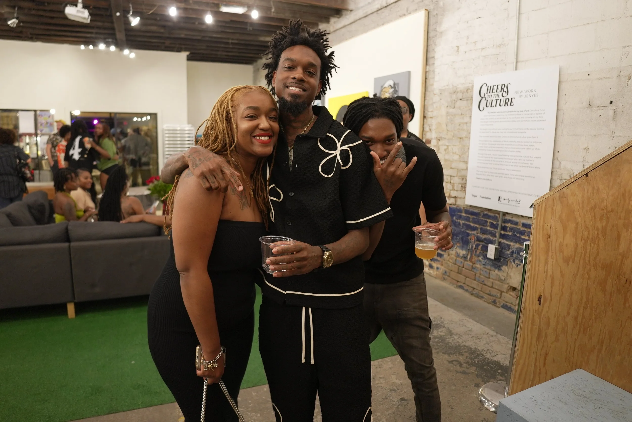 Three people smiling and posing for a photo at an indoor event, two men and a woman, with the woman and one man holding drinks. Other attendees are visible in the background.