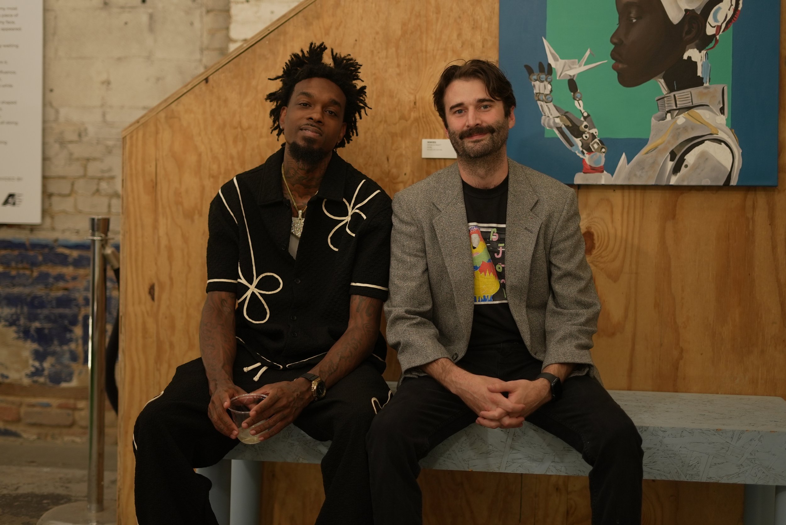 Two men sitting on a bench in an art gallery, with colorful artwork behind them.