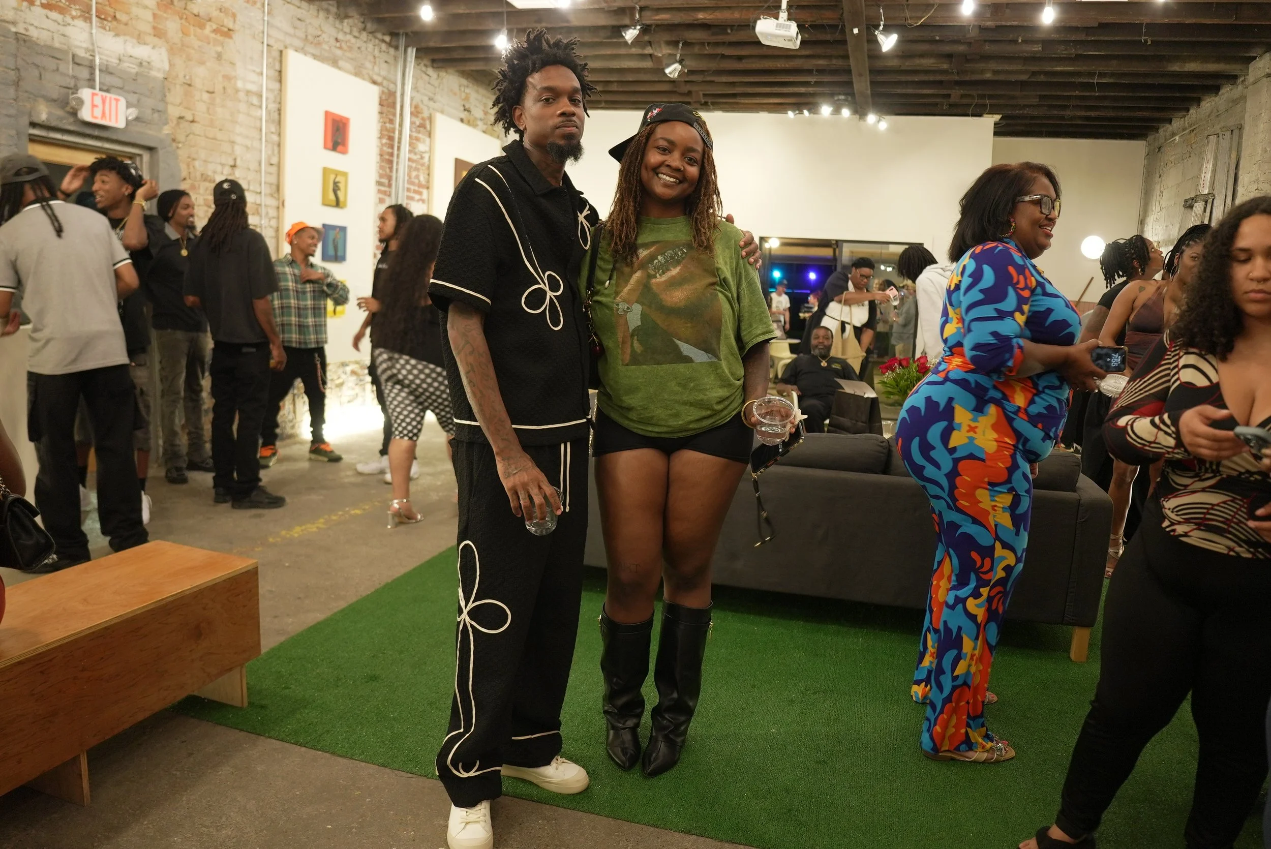 Two people standing in the center of a crowded art gallery or event space, smiling at the camera. The person on the left has short dreadlocks and tattoos, wearing a black outfit with white lines. The person on the right has long dreadlocks, wearing a