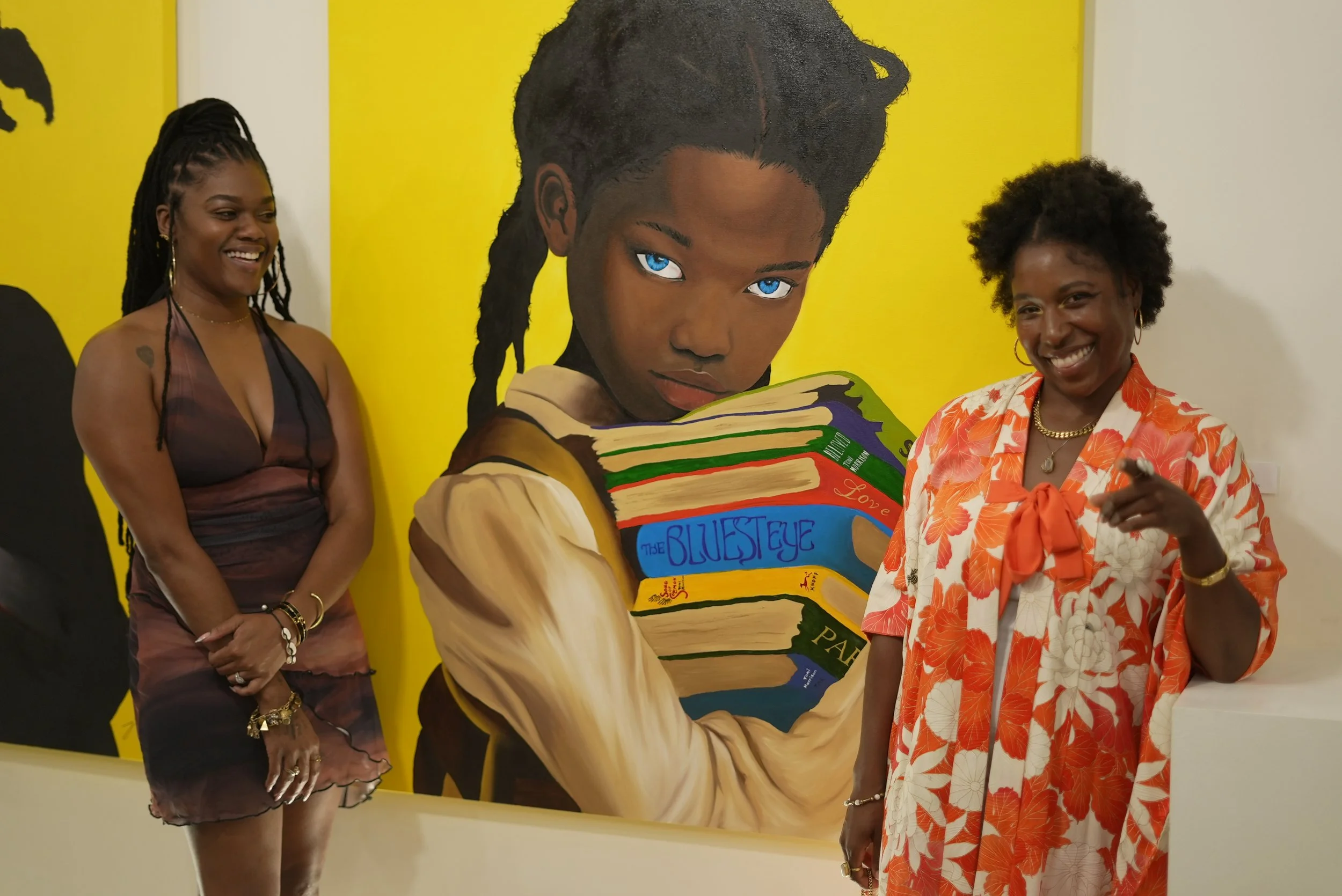 Two women standing beside a large, colorful portrait of a girl with blue eyes reading a book. One woman is wearing a floral-patterned dress, and the other is in a dark dress. Both women are smiling.