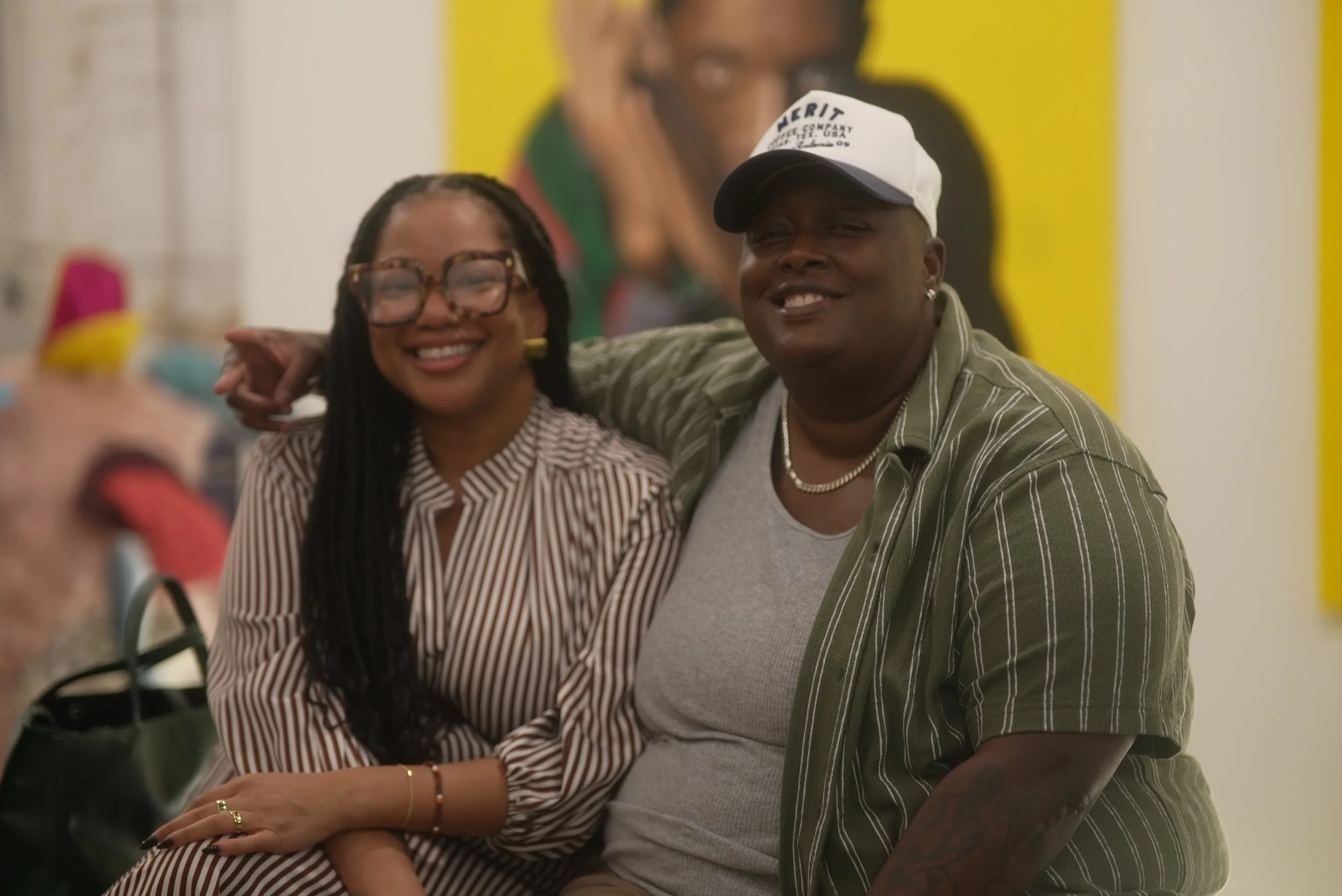 A smiling woman with glasses and long braids, and a smiling man wearing a white cap and a striped shirt, stand together with their arms around each other. They are in an indoor space with a colorful wall and a large portrait in the background.