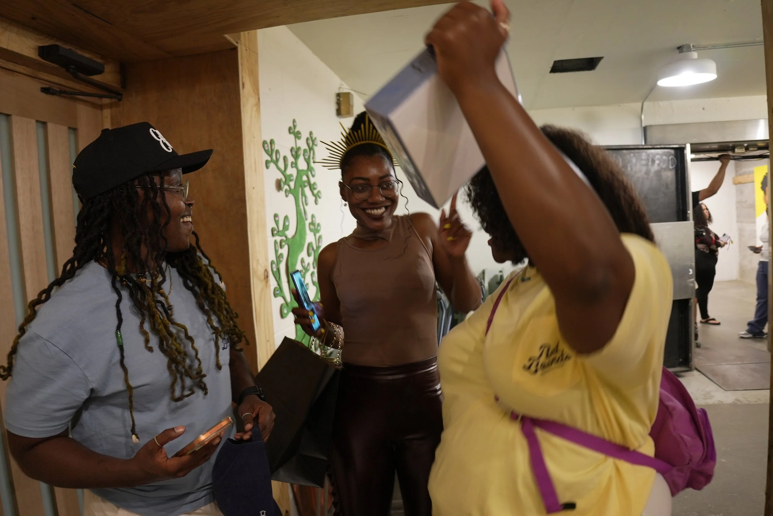 Three women are engaged in conversation in an indoor setting. One woman is wearing a crown and glasses, smiling, holding a phone. The other women are wearing casual clothing, and one of them is raising her arm. The background features a wall with a g