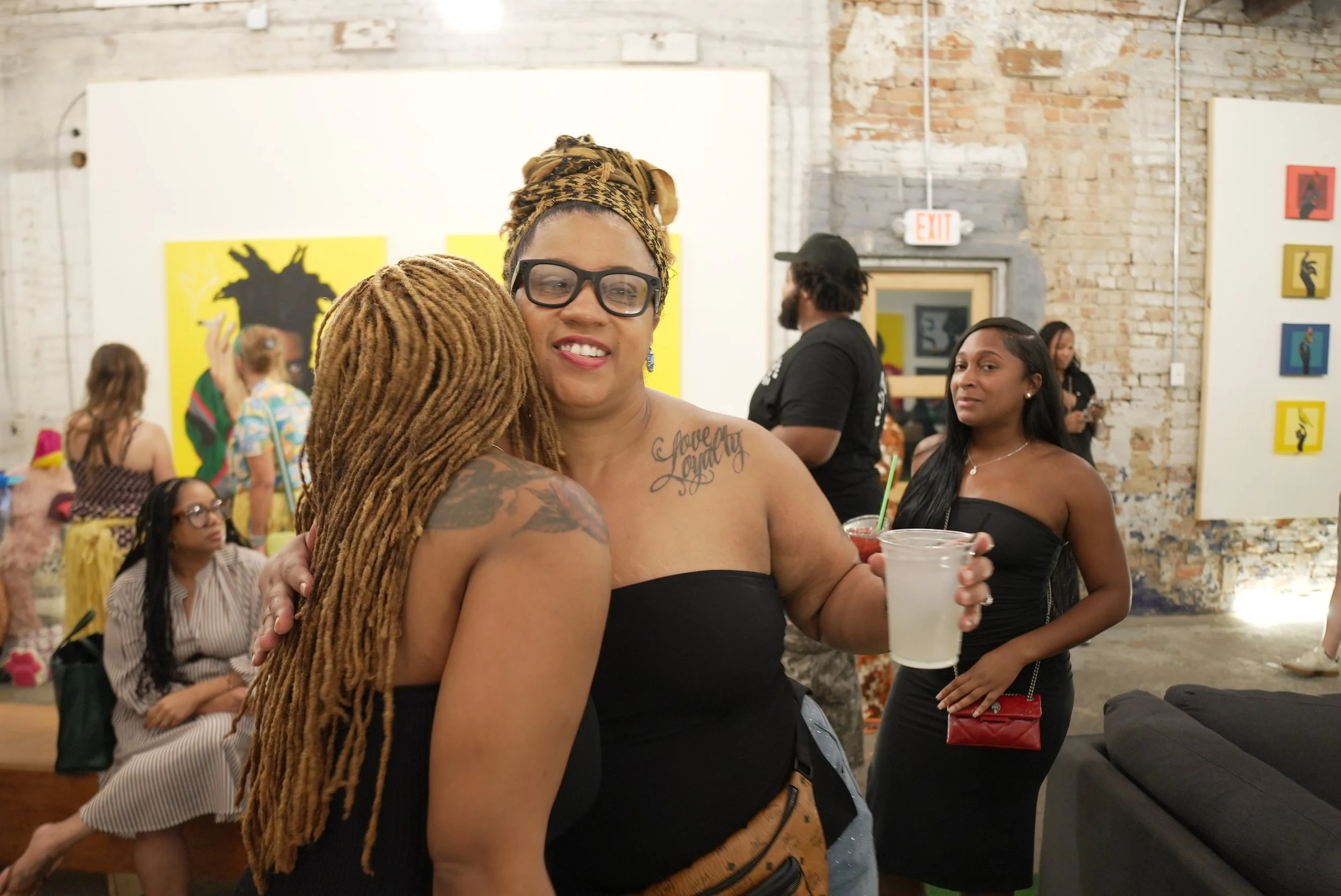 Two women are hugging at a social gathering in an art gallery with exposed brick walls and colorful artwork. One woman has long dreadlocks and the other woman wears glasses and a headwrap. In the background, there are other people, including a woman 