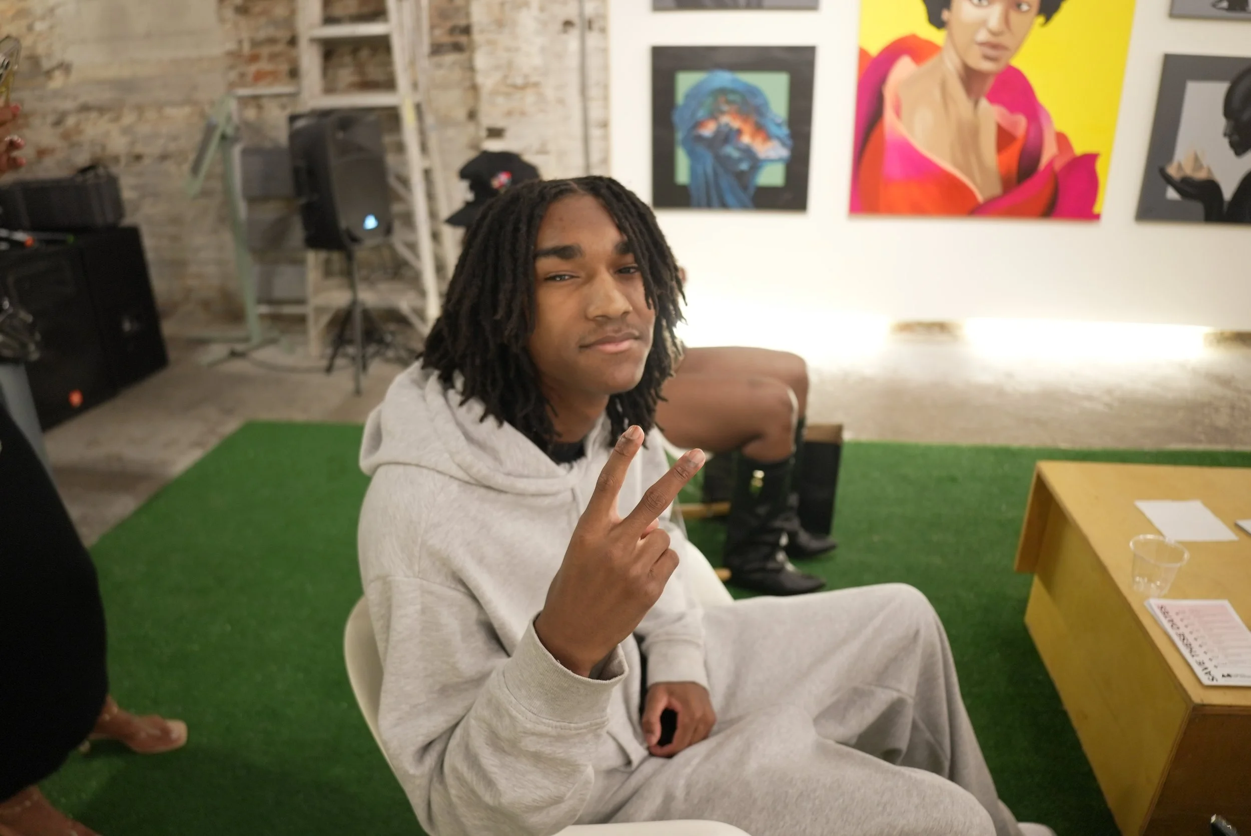 A young man with dreadlocks and a white hoodie sitting indoors on a white chair, making a peace sign with his right hand. Behind him, there are colorful artworks on the wall, including a woman with short hair in bright colors. The setting appears to 