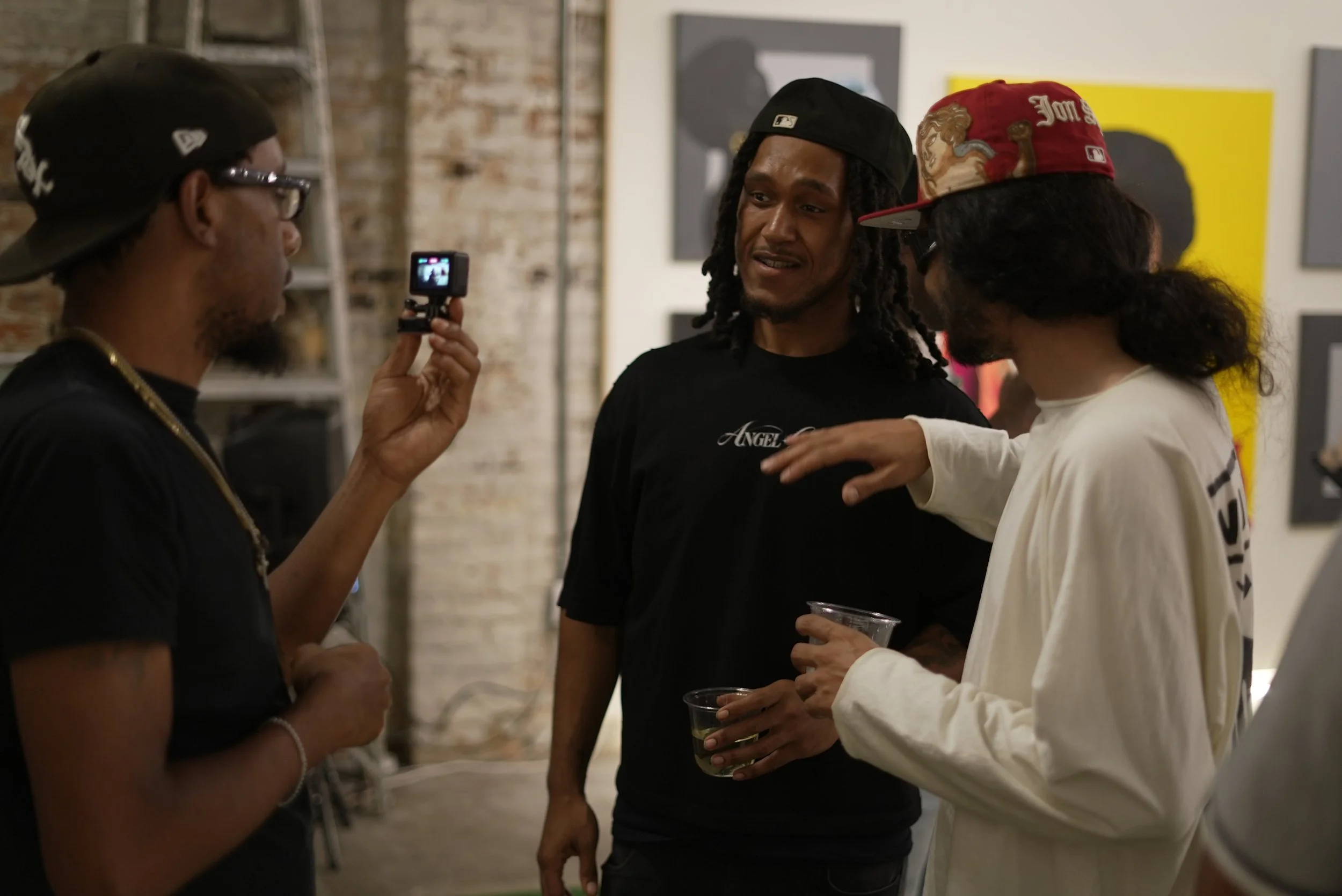 Four people are conversing in an art gallery. The man on the left wears a black cap and glasses, holding a small camera. The man in the middle has dreadlocks, a black cap, and a black T-shirt. The woman on the right has long dark hair, wearing a whit