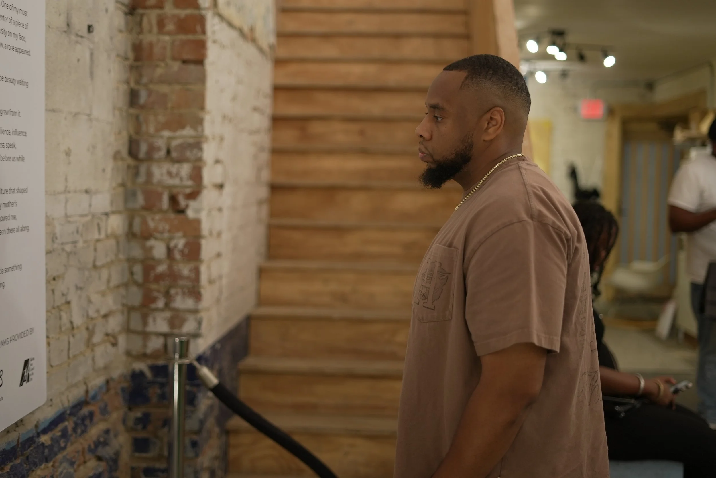 A young man with a beard and short haircut looking at a display or artwork on a brick wall in an indoor gallery or museum space. He is wearing a brown shirt and a gold chain.