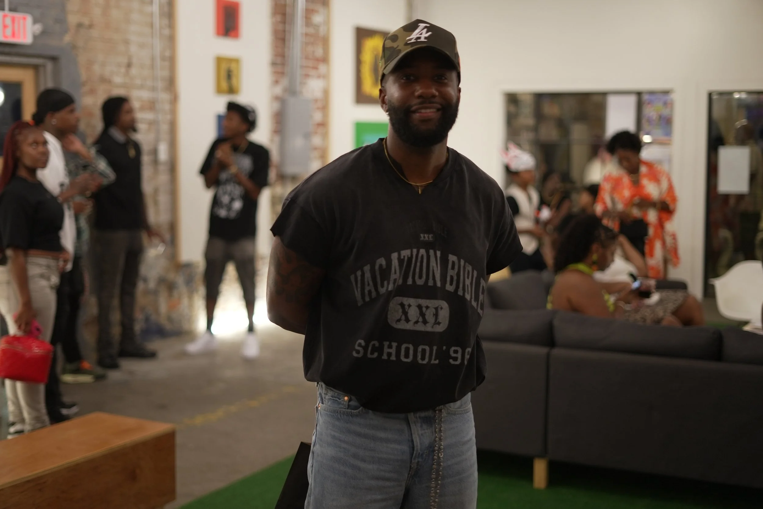 A man with a beard wearing a black LA cap and a black T-shirt, standing in an art gallery or event space, smiling at the camera. In the background, there are several women, some wearing black, standing and sitting, with art pieces on the wall behind 