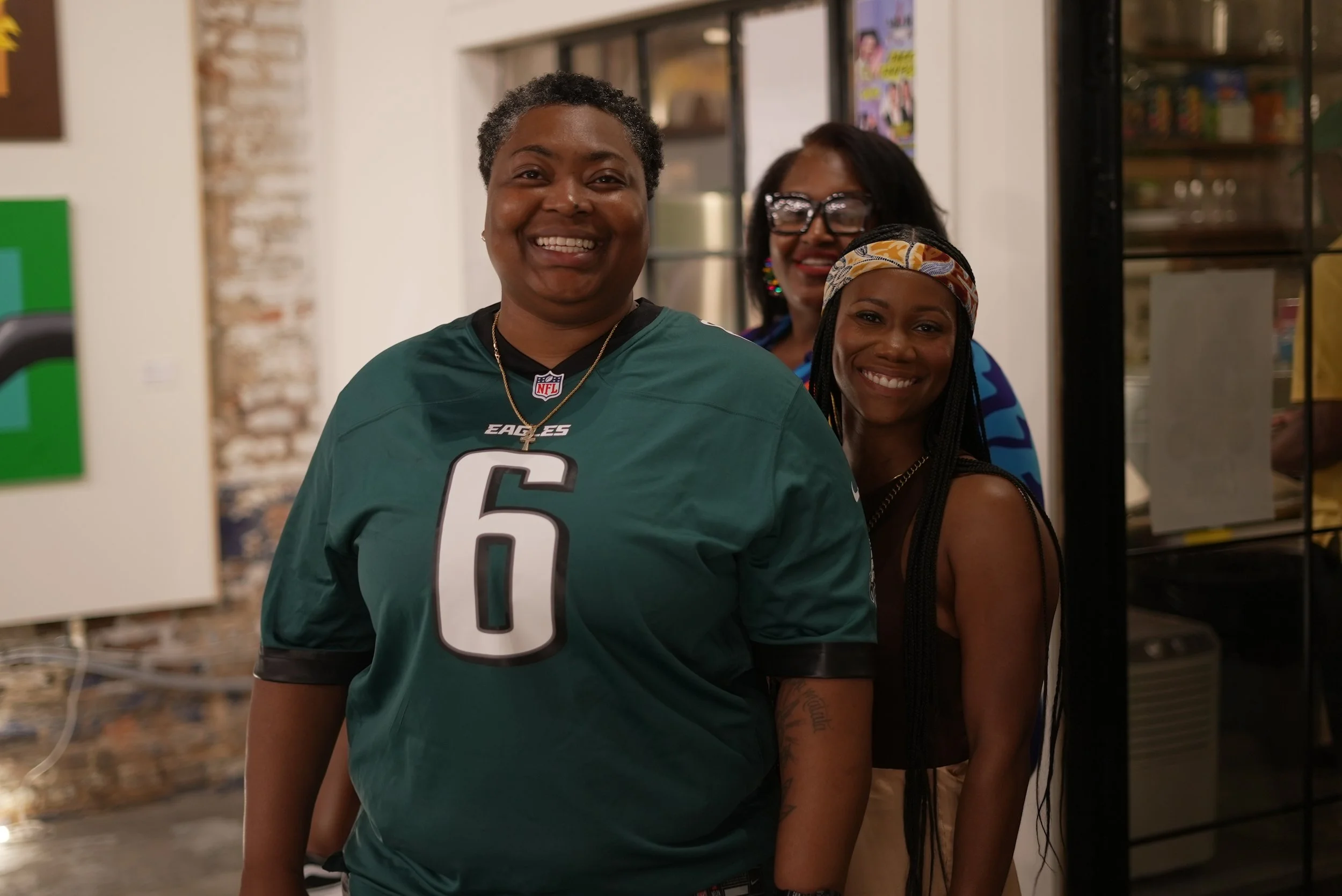 Three women smiling and standing together indoors. One woman in the foreground wears a Philadelphia Eagles football jersey. Two women are partially visible behind her, smiling.