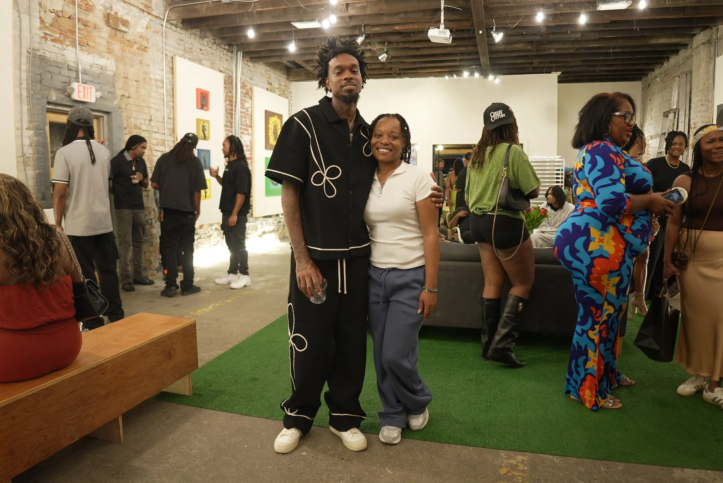 Two people standing together at an art gallery event, smiling at the camera. The gallery has exposed brick walls, colorful artwork, and a group of people mingling in the background.