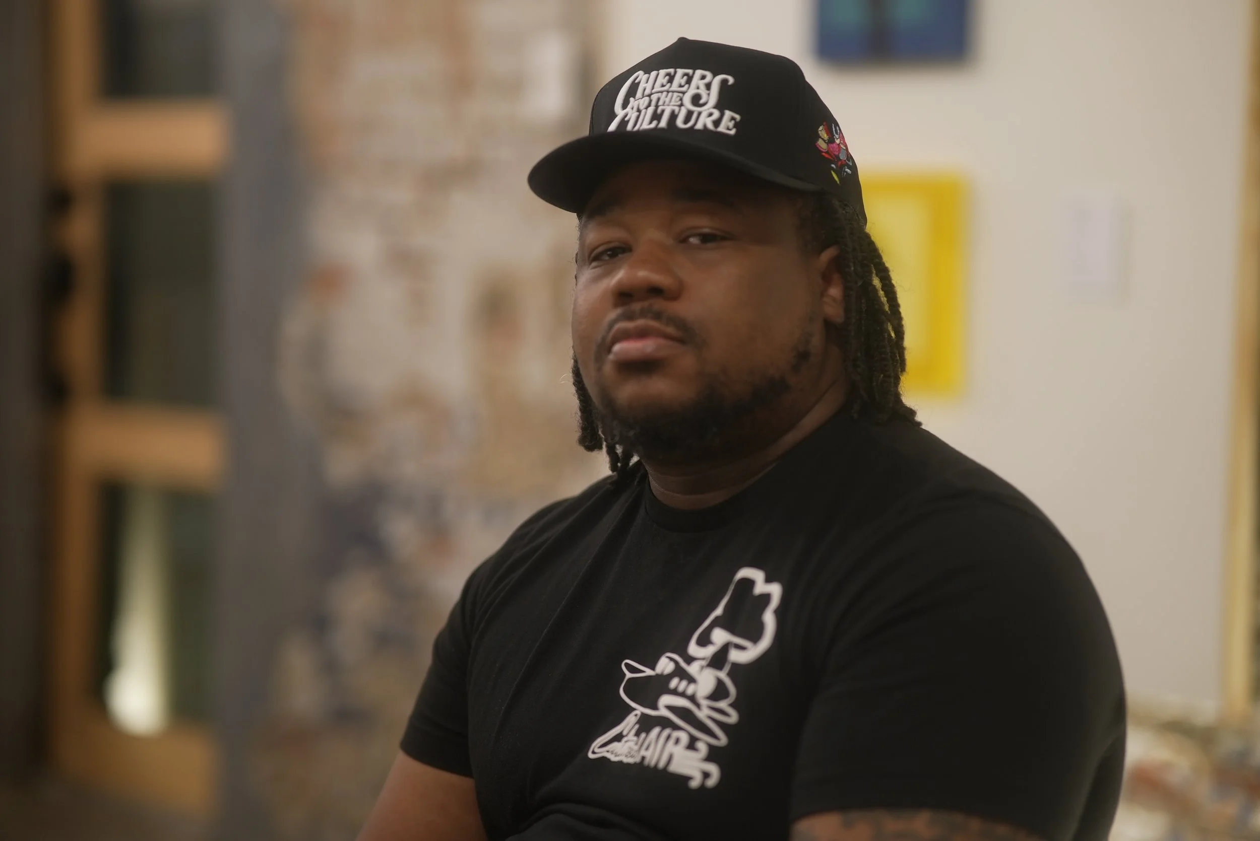 A young man with dreadlocks wearing a black cap with the text "Cheer Quotes Culture" and a black T-shirt with a graphic design, standing indoors with a blurred background of a brick wall and framed artworks.