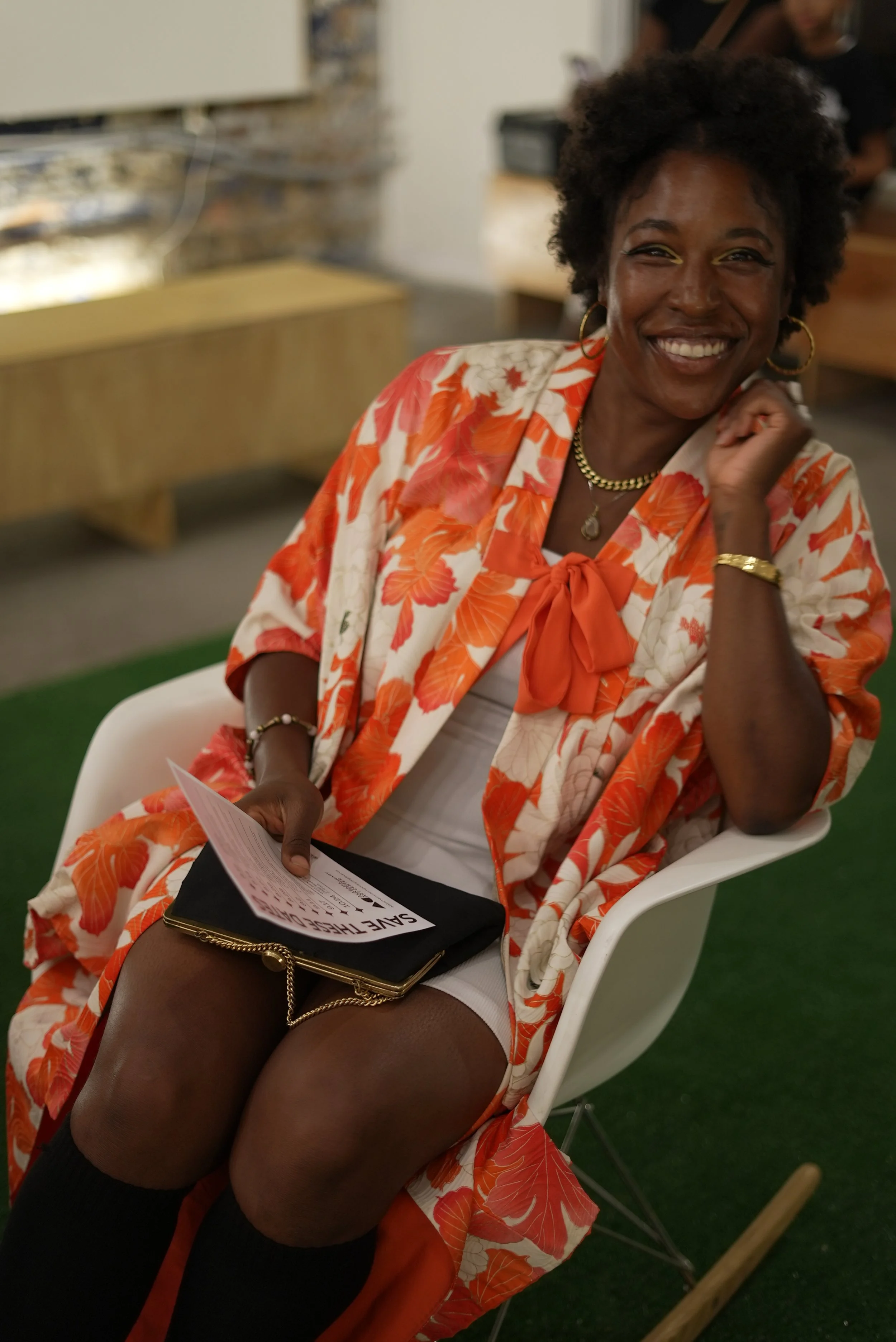 A smiling woman with curly hair, wearing a floral orange and white jacket, gold jewelry, and black stockings, sitting on a white chair indoors.