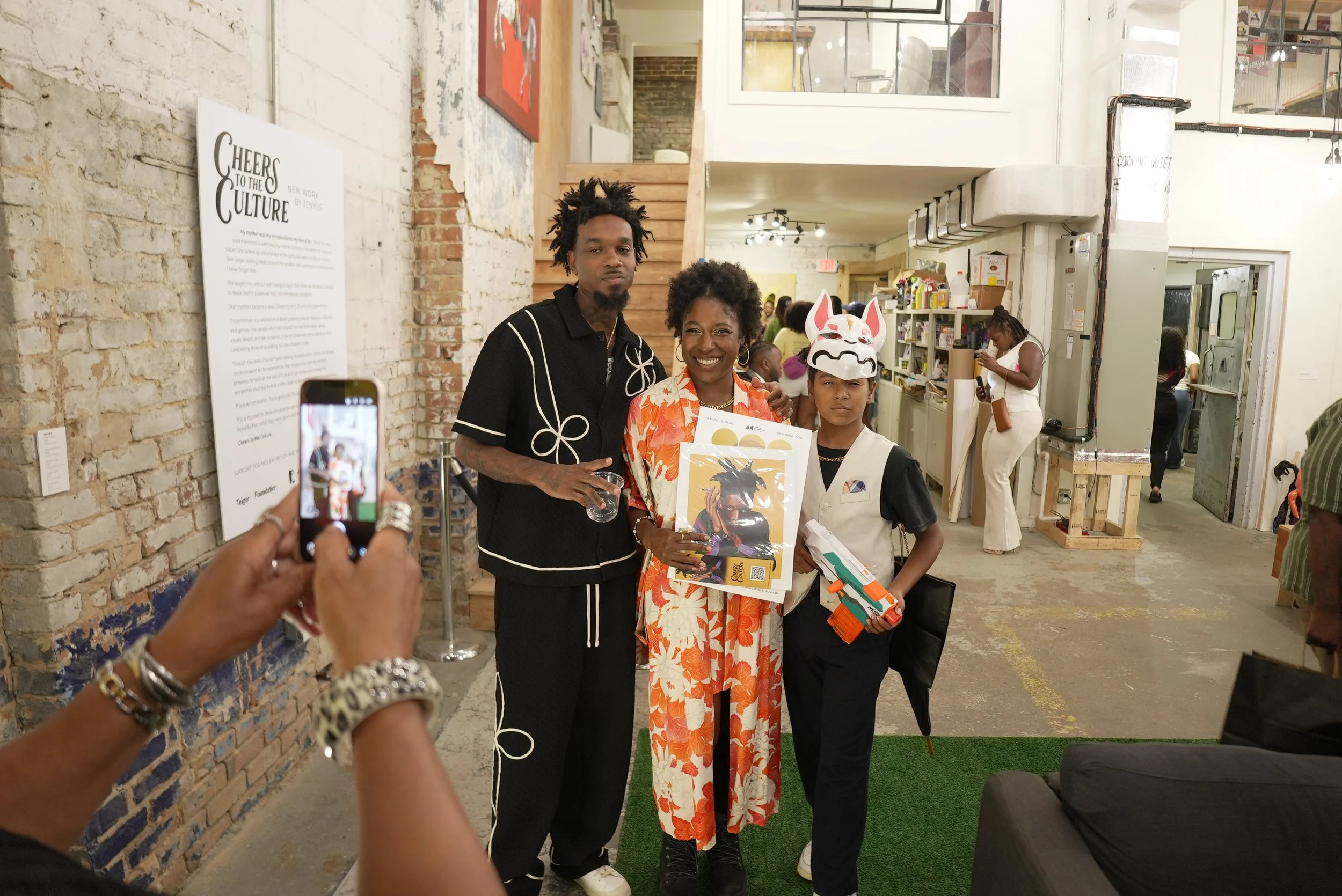 Three people standing together inside a gallery or event space, posing for a photo. One woman is holding a colorful flyer, while a young girl holds a toy gun and is wearing a traditional fox mask. A man with dreadlocks stands beside them. The backgro
