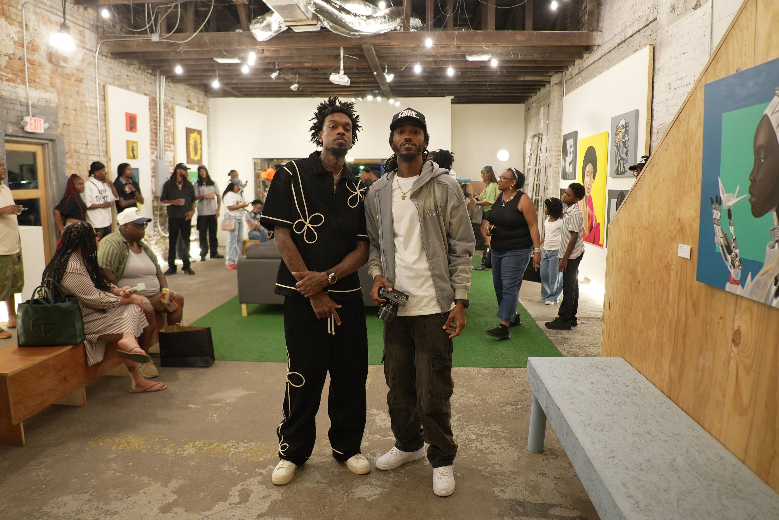 Two young men standing together in an art gallery, surrounded by other visitors and colorful artwork on the walls.