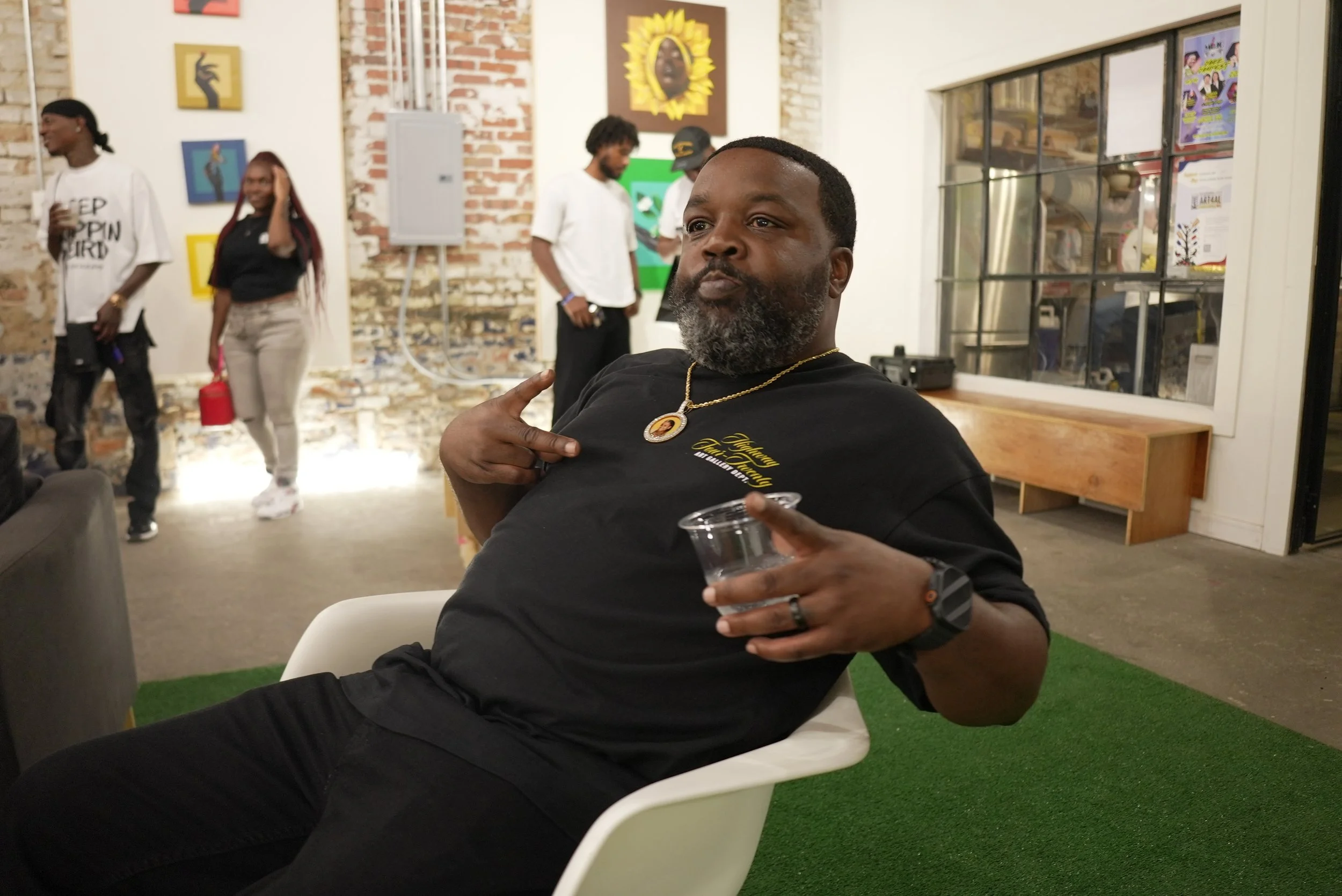 A man with a beard and short hair, wearing a black T-shirt with yellow writing and a gold chain with a pendant, is sitting in a modern art gallery or event space. He is holding a plastic cup in one hand and pointing towards himself with the other. Be