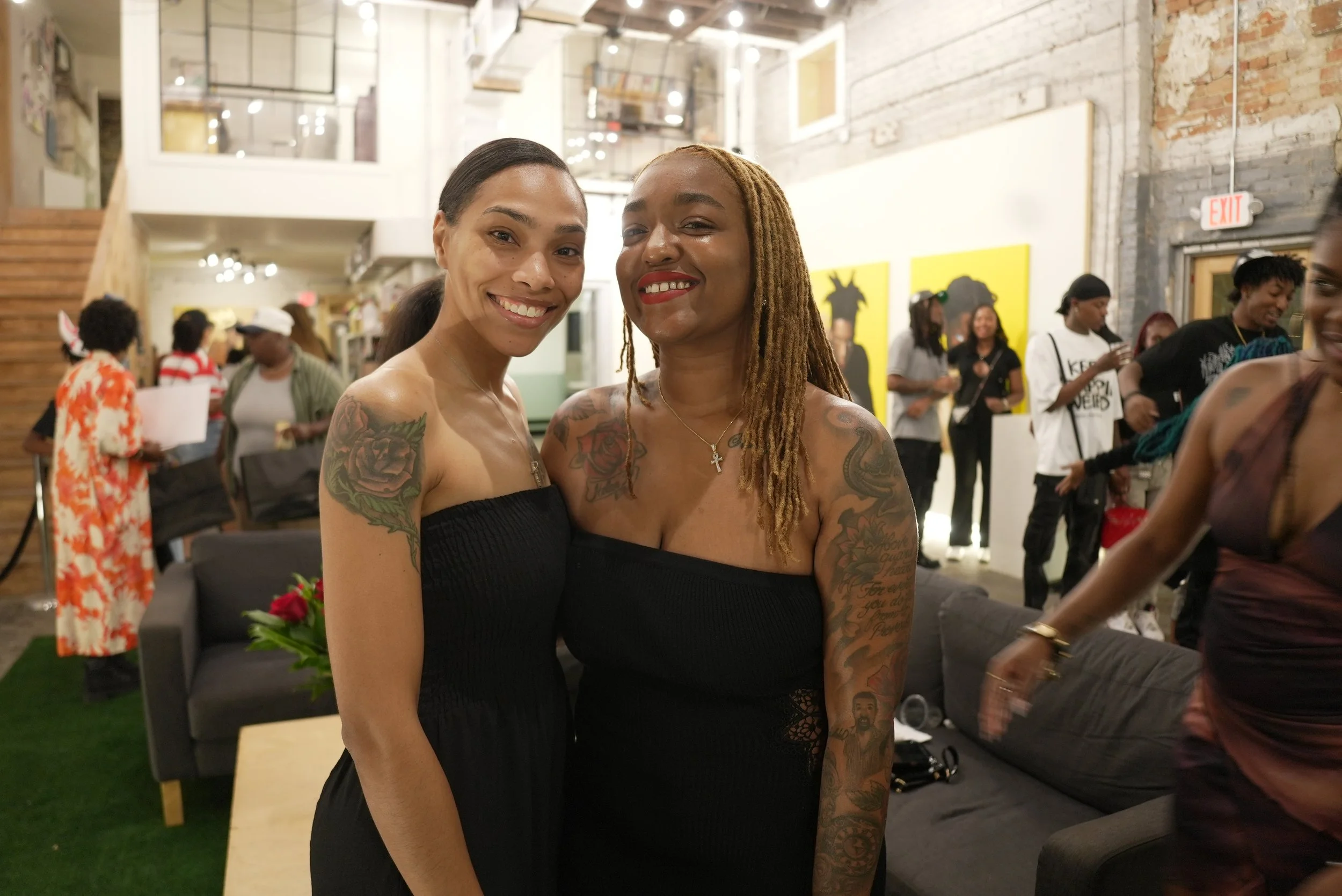 Two women smiling and looking at the camera at an indoor event with many people in the background. One woman has dark hair and tattoos on her arm, and the other has blonde dreadlocks and tattoos on her shoulder.