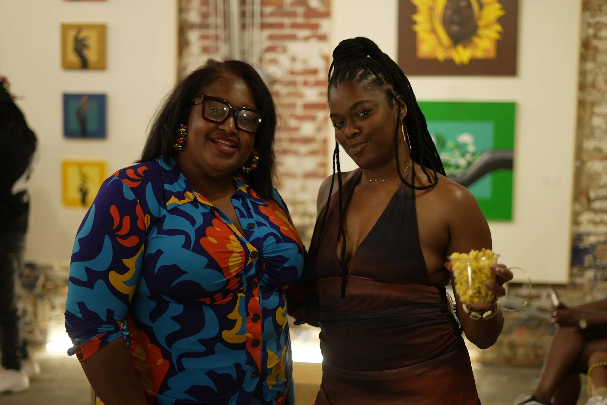 Two women standing close together at an art gallery, smiling. One woman wears a colorful, patterned blouse and glasses, the other is in a sleeveless, dark top with dreadlocks, holding a bowl of popcorn.