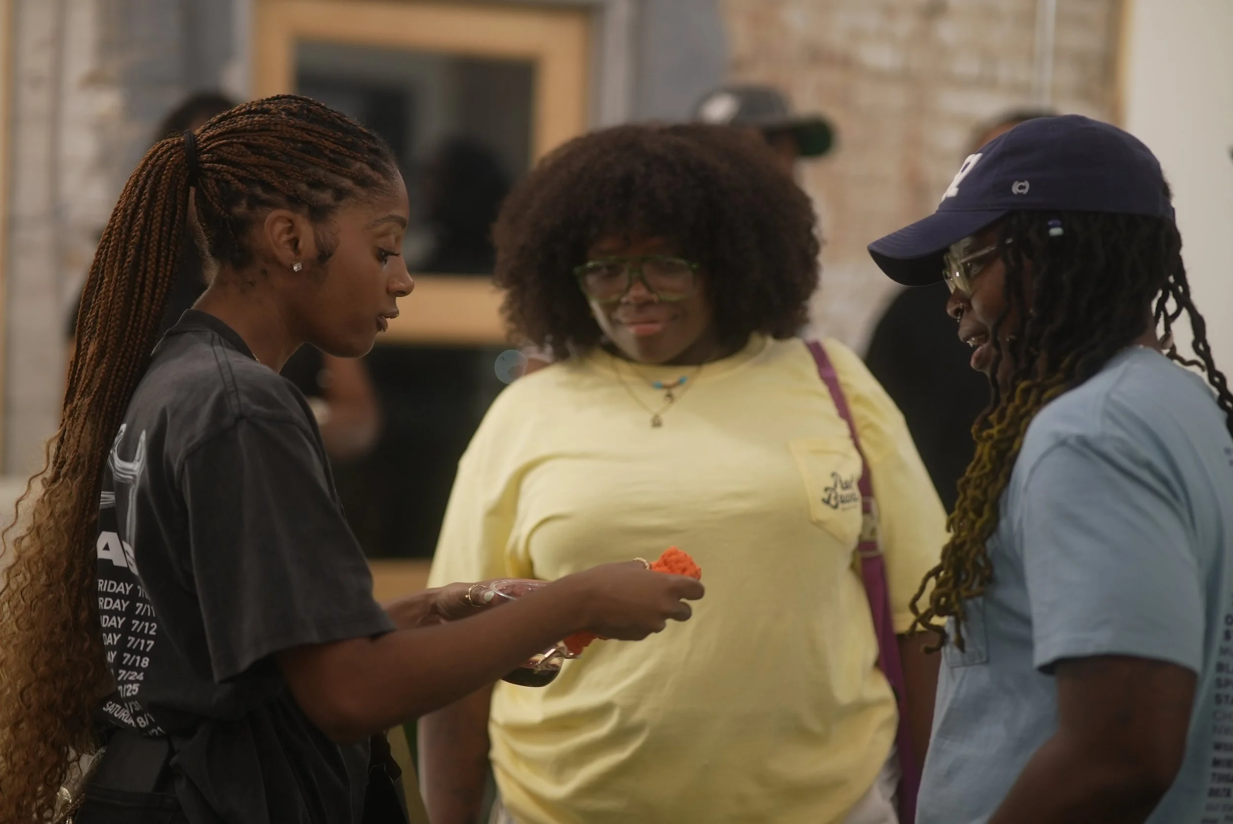 Three women engaged in conversation in an indoor setting. One woman has long braided hair and is wearing a black shirt. The second woman has curly hair, glasses, and is wearing a yellow shirt. The third woman has dreadlocks, a backwards baseball cap,