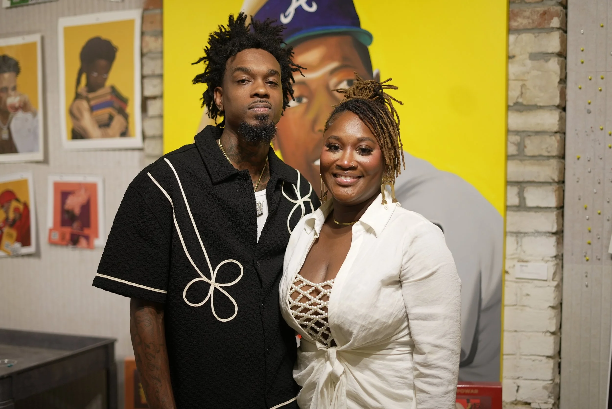 A man and a woman standing side by side indoors, with art and photographs displayed on the wall behind them. The man has dark dreadlocked hair, a beard, and tattoos, wearing a black shirt with white piping. The woman has short, styled dreadlocks, wearing a white blazer over a decorative inner top, and large earrings, smiling at the camera.