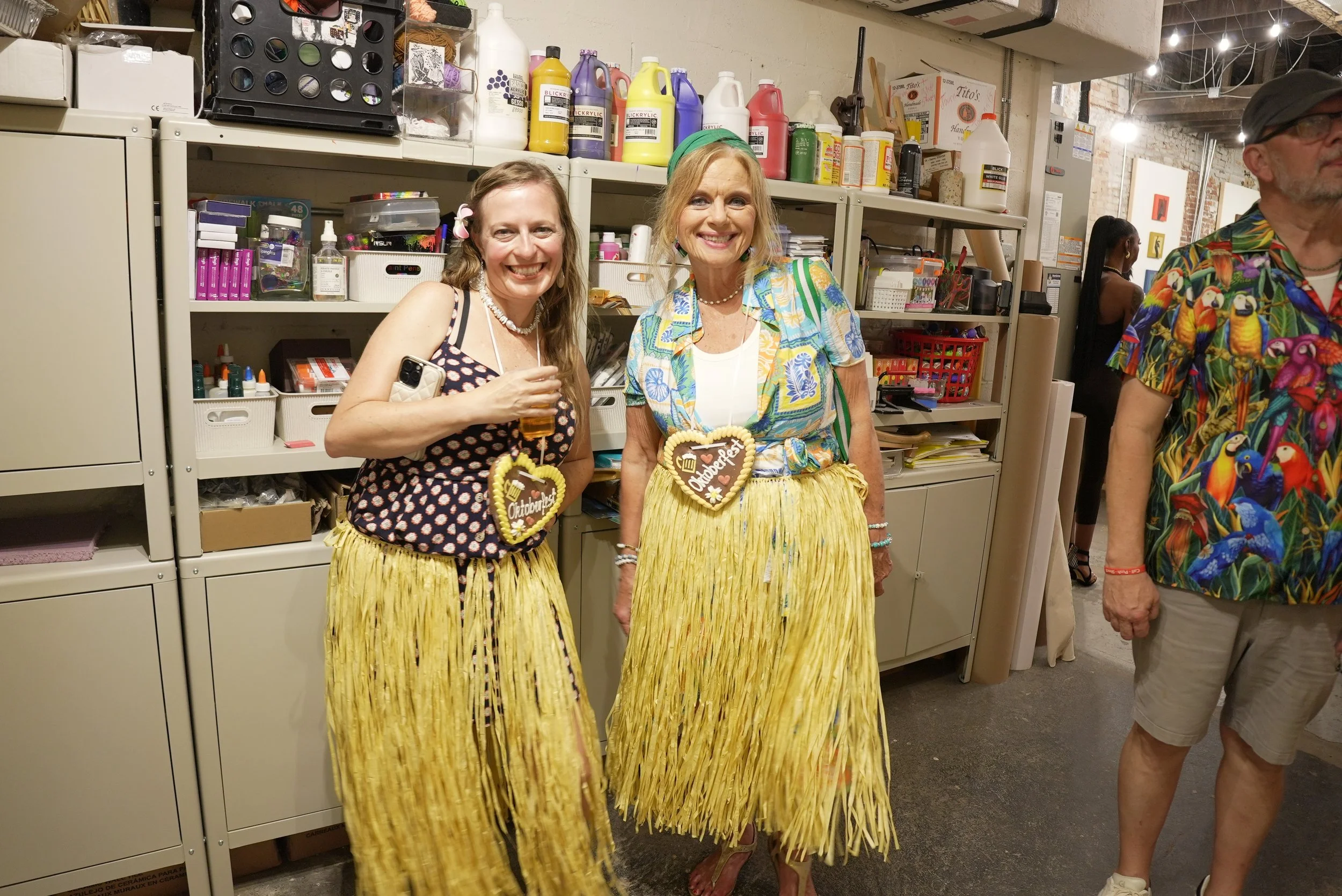 Two smiling women dressed in black and floral patterned tops and yellow grass skirts, standing in what appears to be an arts and crafts studio, with shelves of supplies behind them.