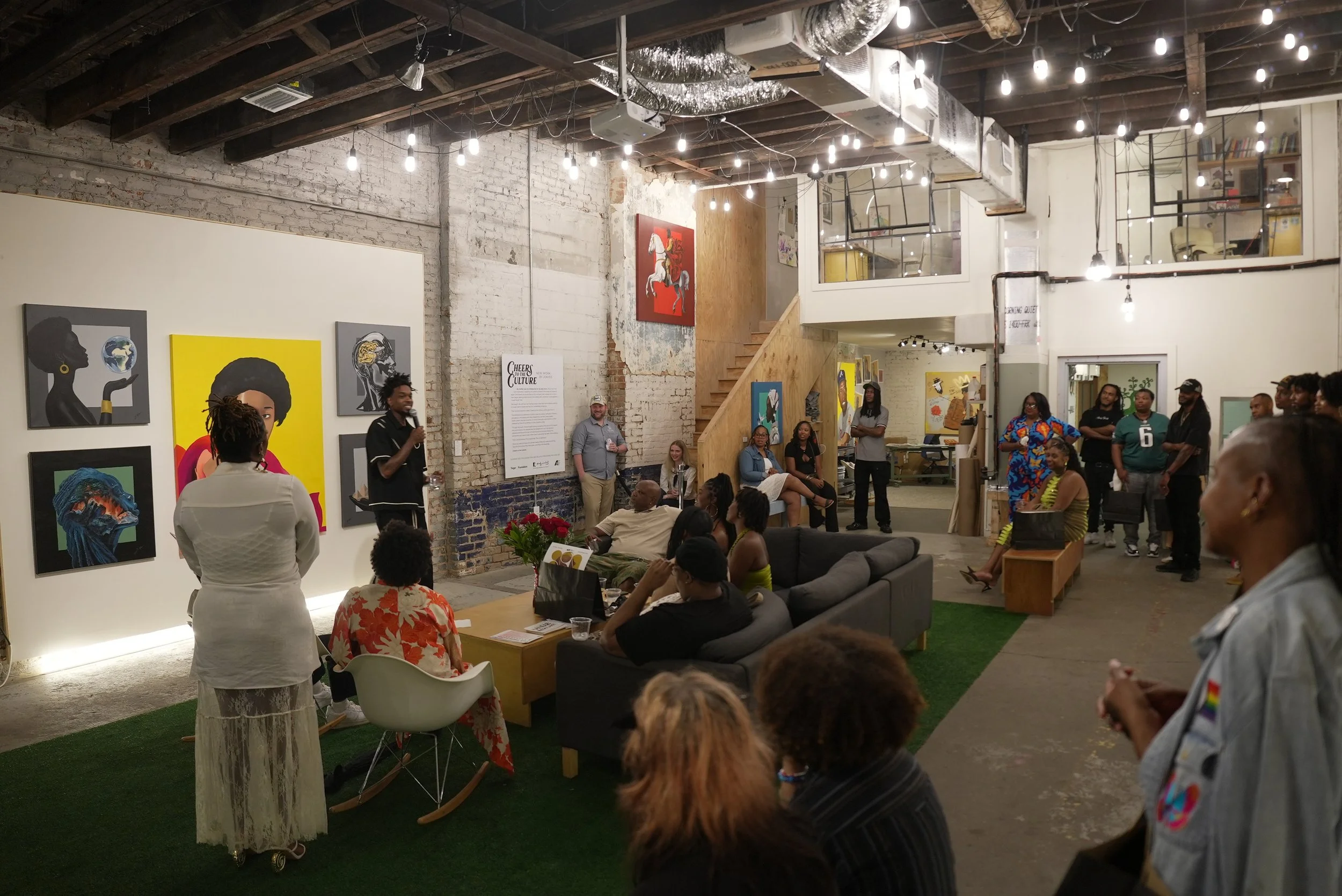 People gathered in an art gallery with paintings on the wall and a speaker holding a microphone.