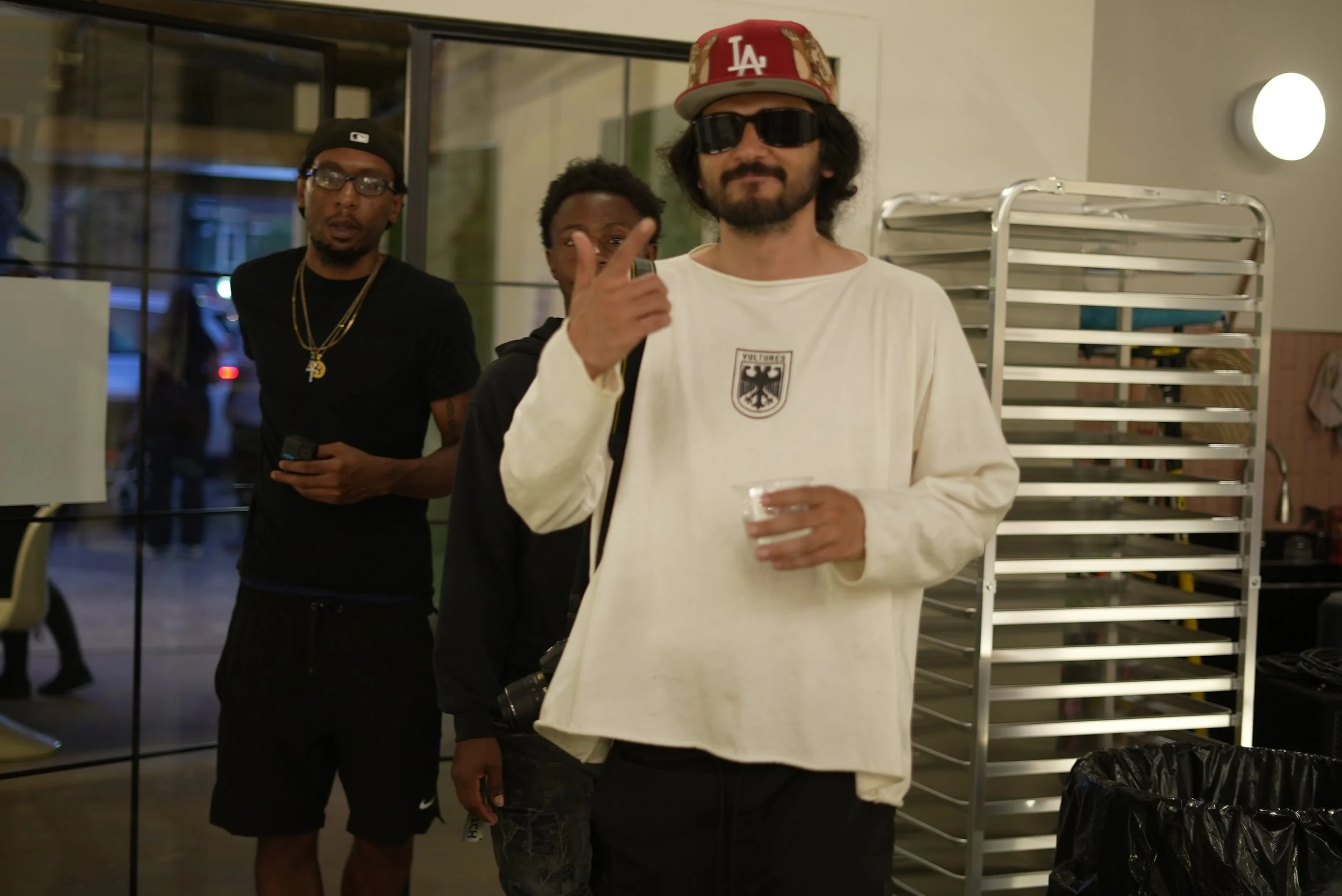 Three men standing indoors, one in the foreground wearing sunglasses, a red LA baseball cap, a white T-shirt with a German eagle emblem, and holding a clear plastic cup. Two men are behind him, one wearing glasses, a black cap, and a black shirt, and